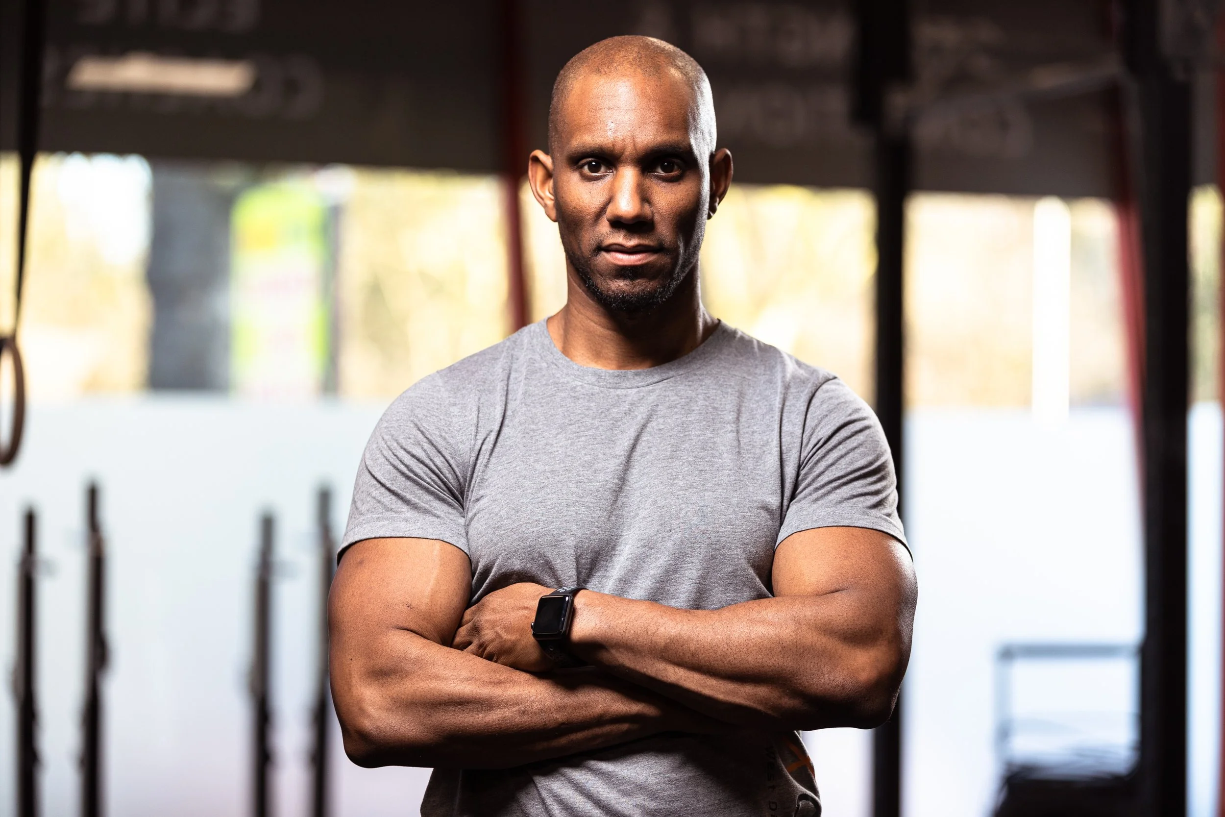 A man with a bald head and wearing a gray t-shirt stands with arms crossed in a gym, looking at the camera.