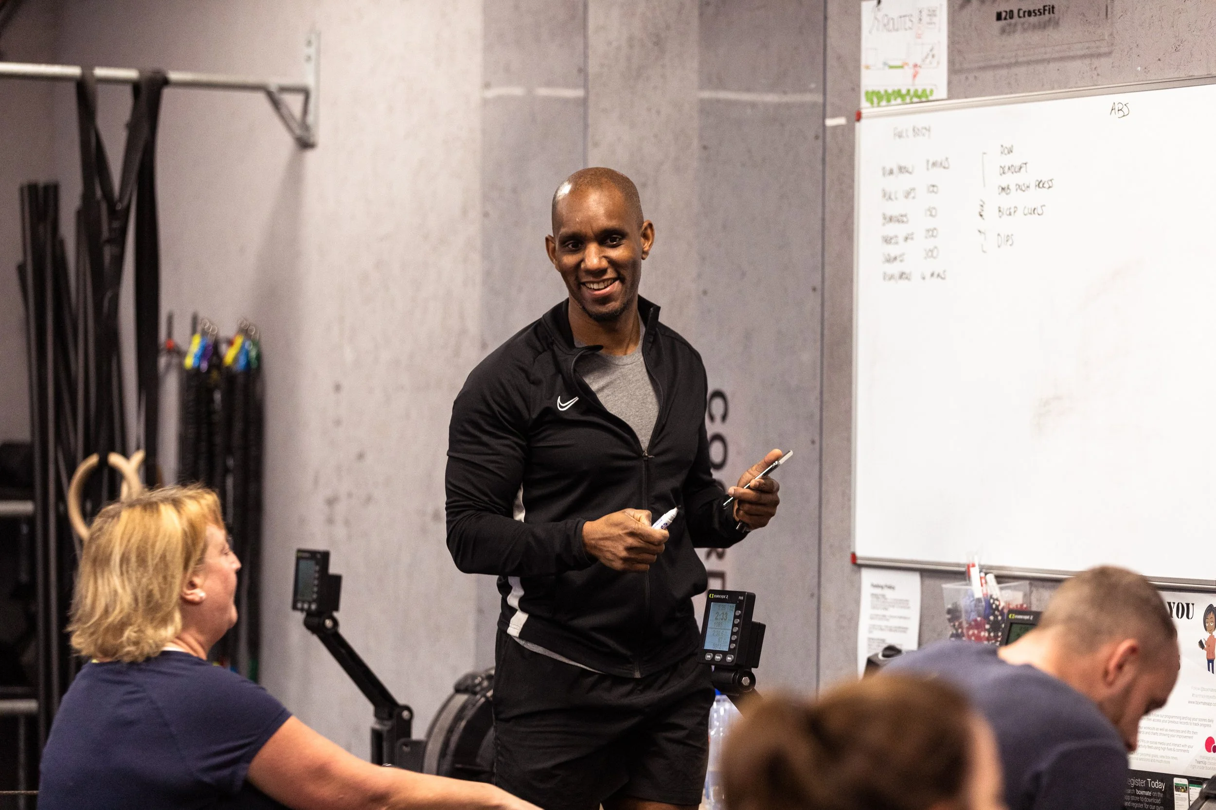 A man in a black jacket and gray shirt smiling in a gym or fitness class, holding a marker, standing near a whiteboard with handwritten notes, with other people sitting at a table in front of him.