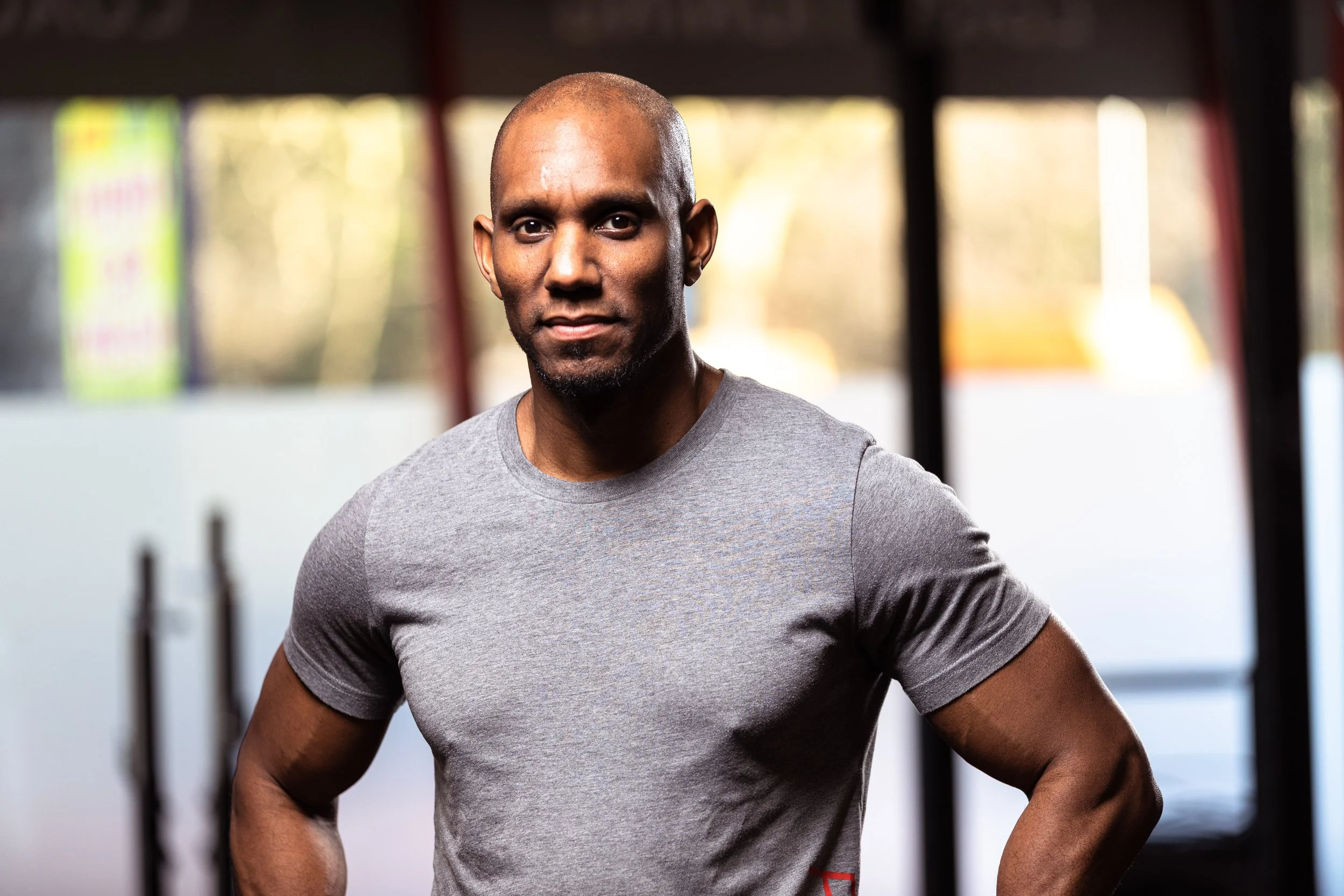 A muscular man with a shaved head and a serious expression is standing in a gym wearing a gray t-shirt.