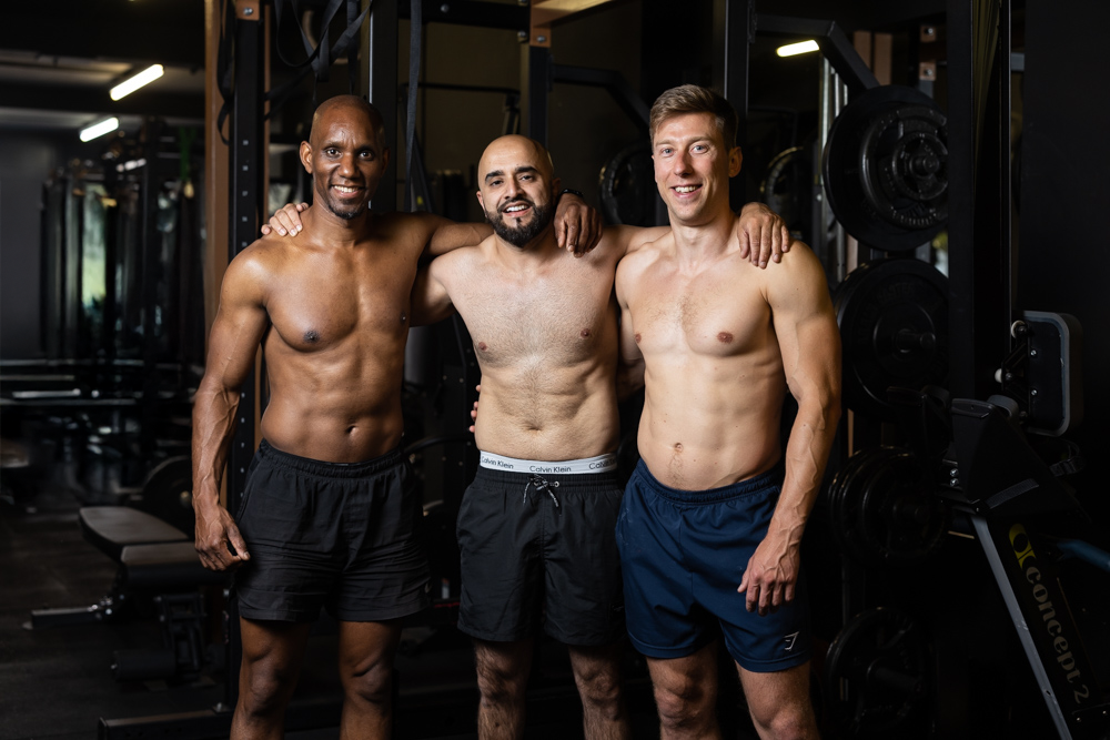Three shirtless men with athletic physiques smiling and standing together in a gym, arms around each other's shoulders.