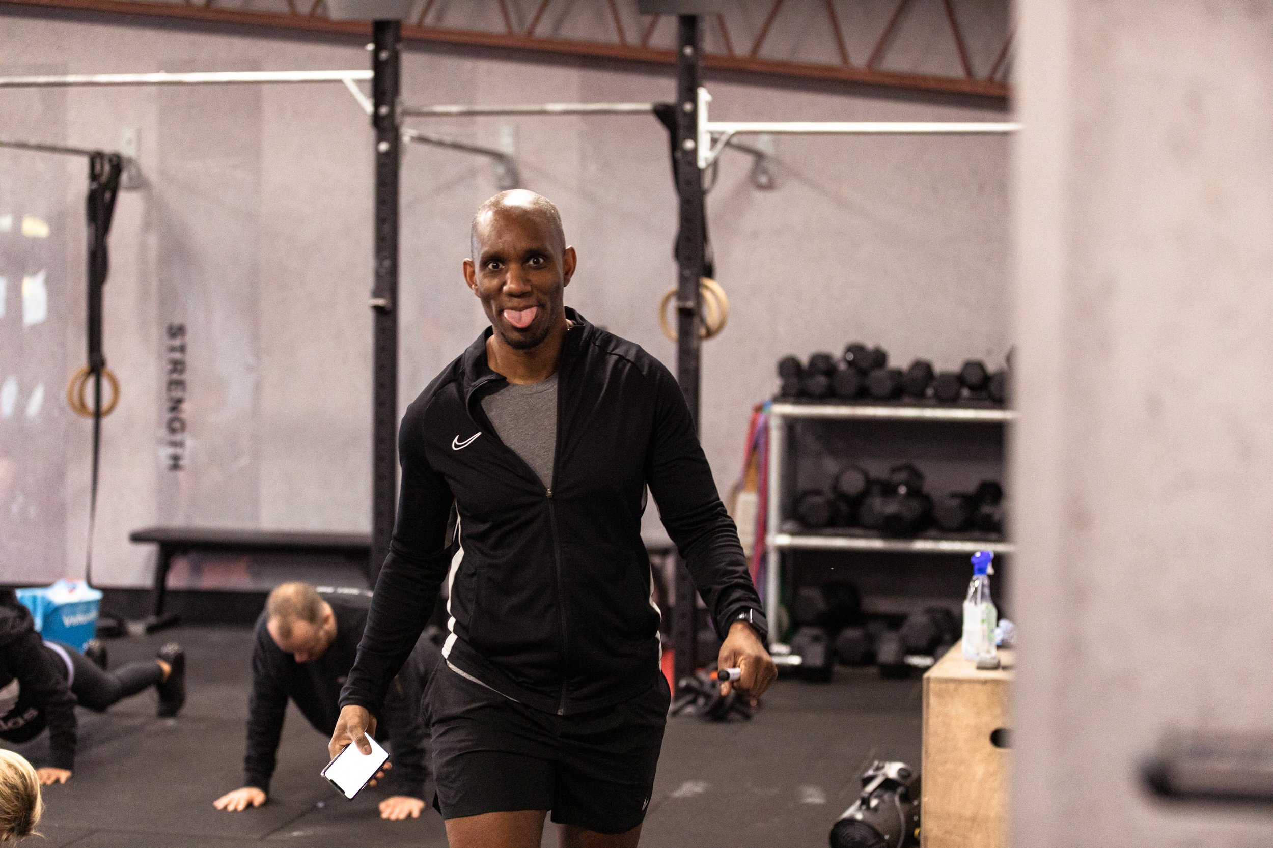 A man in a black athletic jacket and shorts making a funny face with his tongue out in a gym, with people doing push-ups in the background and gym equipment around.