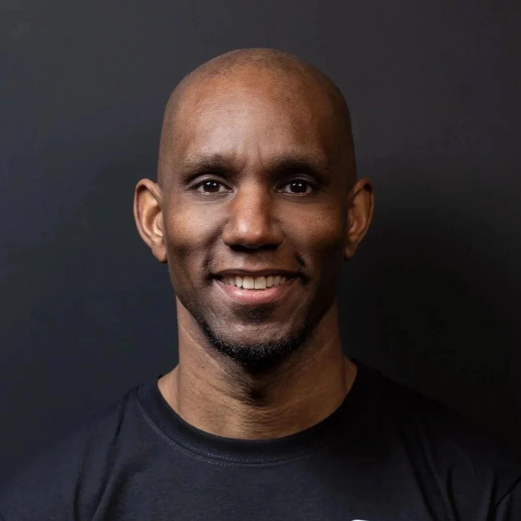 Headshot of a smiling man with a shaved head and a goatee, wearing a black shirt, against a dark background.
