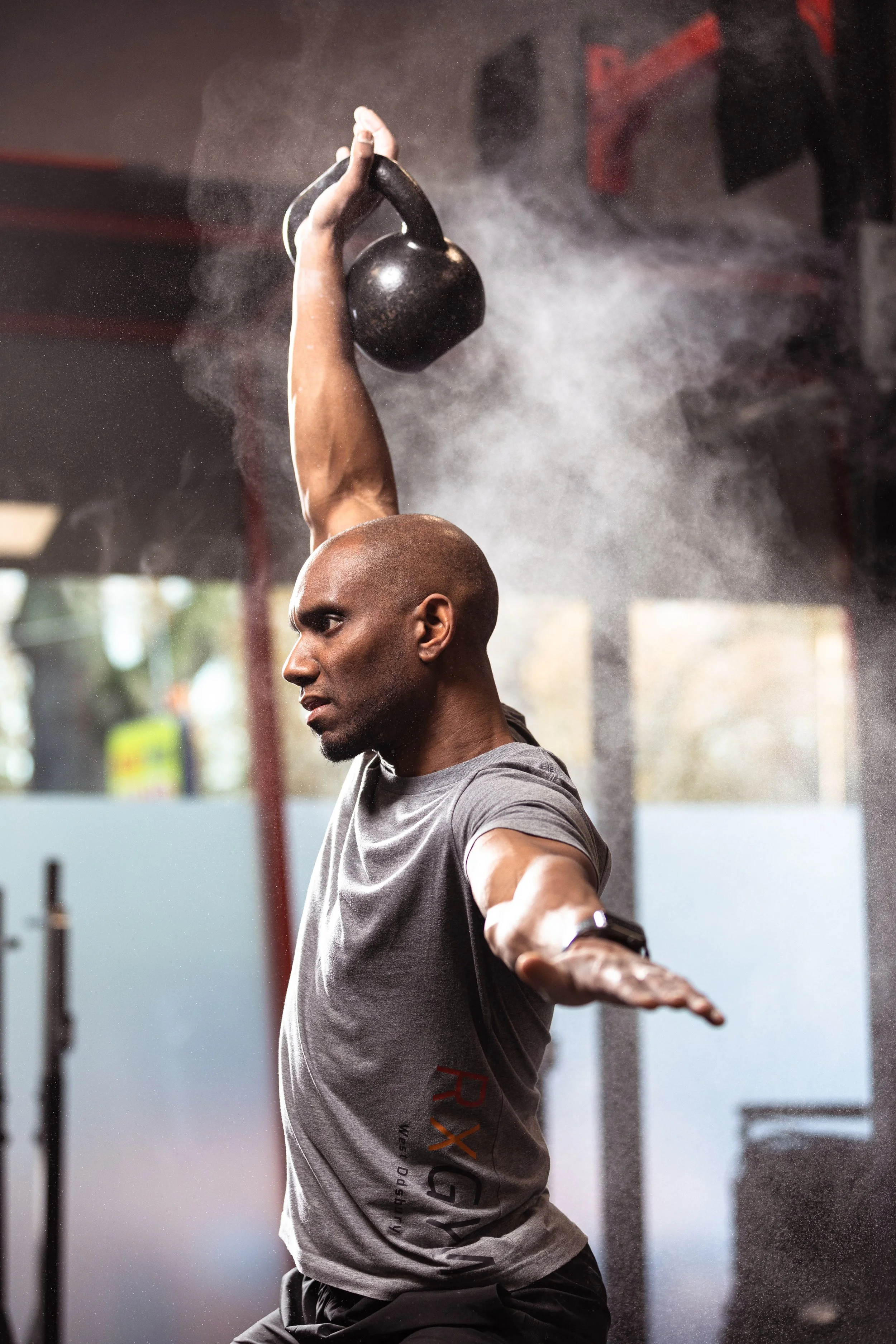 A man with a shaved head lifting a kettlebell overhead in a gym, with chalk dust in the air.