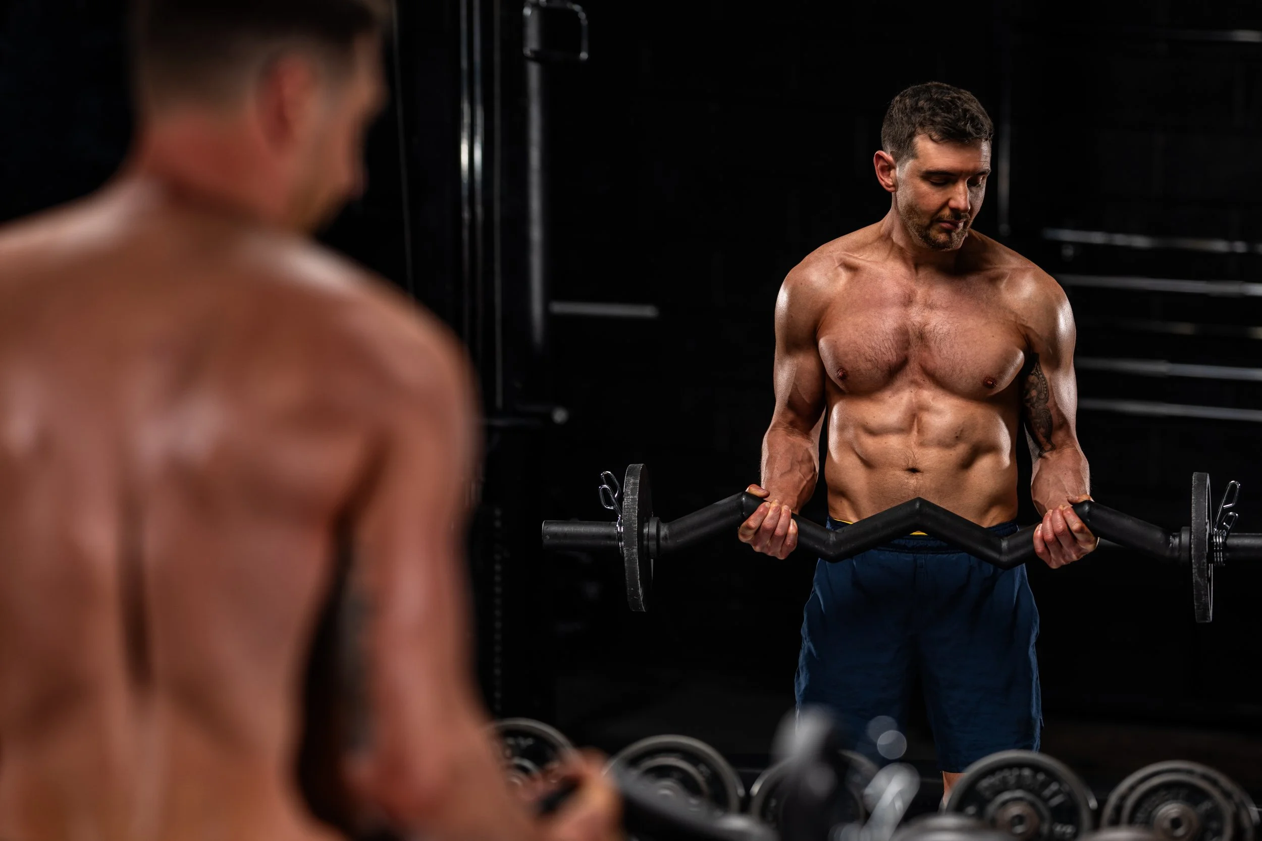 A shirtless man with a muscular build lifting a barbell in a dark gym.
