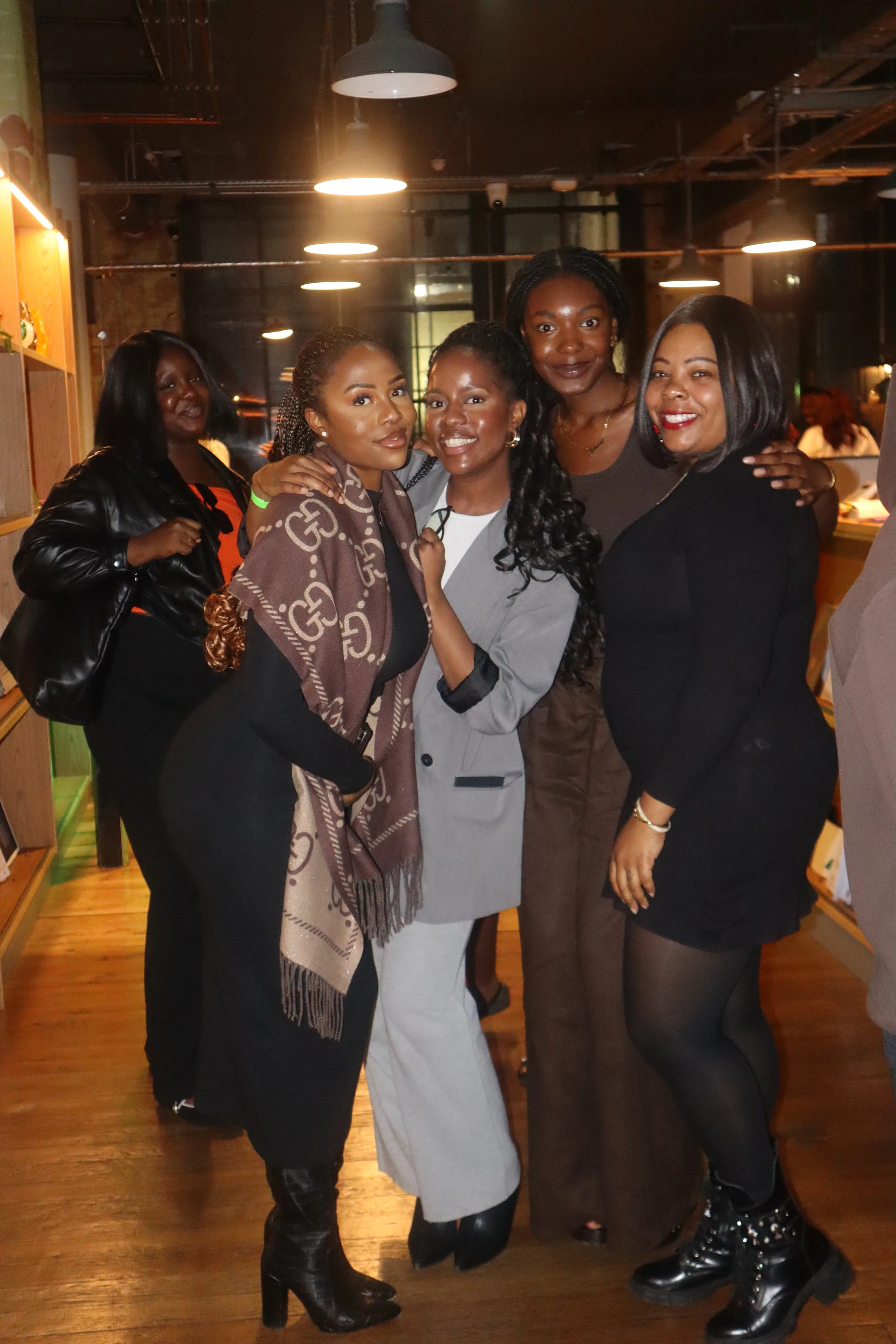 Group of five women in stylish outfits posing together indoors with warm lighting.