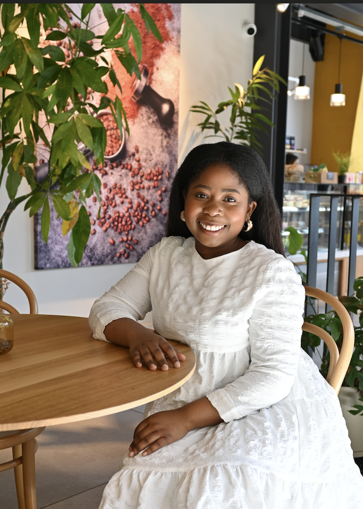 A young girl with dark hair and a white dress sitting at a wooden table in a cafe, smiling, surrounded by green plants and some artwork on the wall.