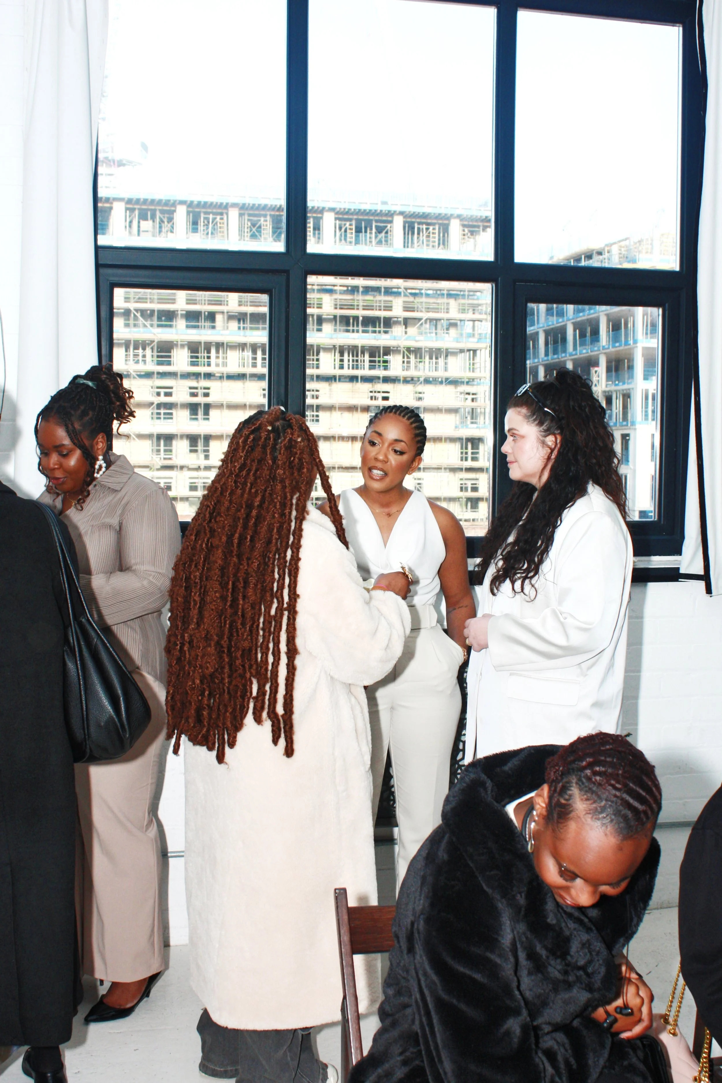 Group of women conversing indoors near large windows with city buildings outside.