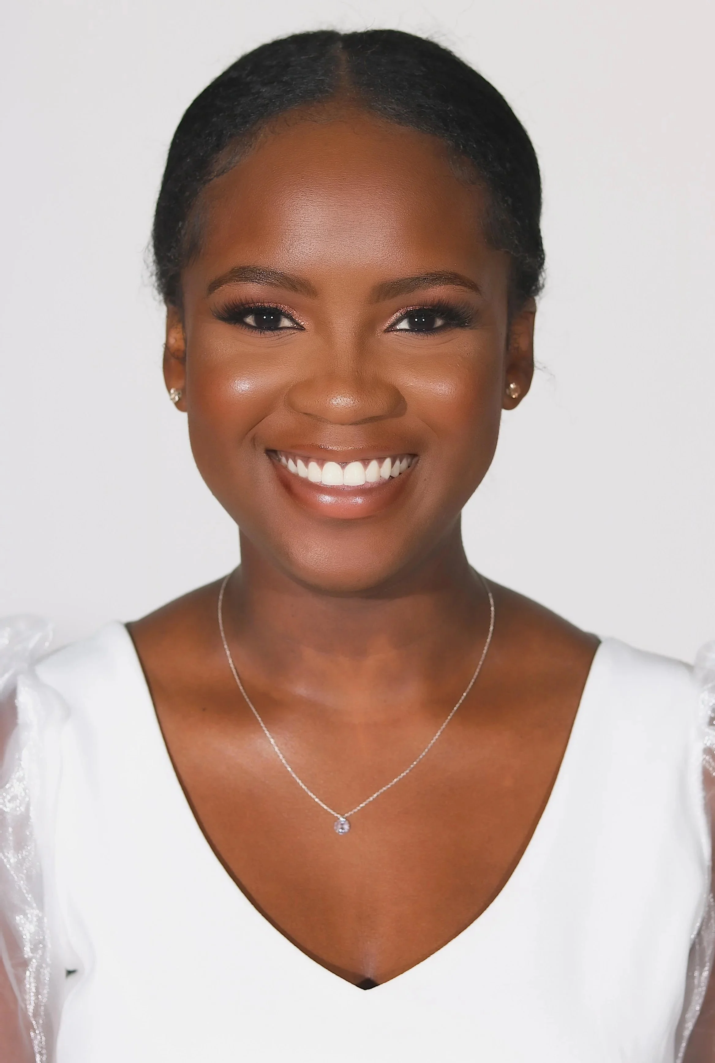 A woman with dark skin and natural hair styled back, smiling and wearing a white top with puffed sleeves and a silver necklace with a pendant, posed against a plain white background.