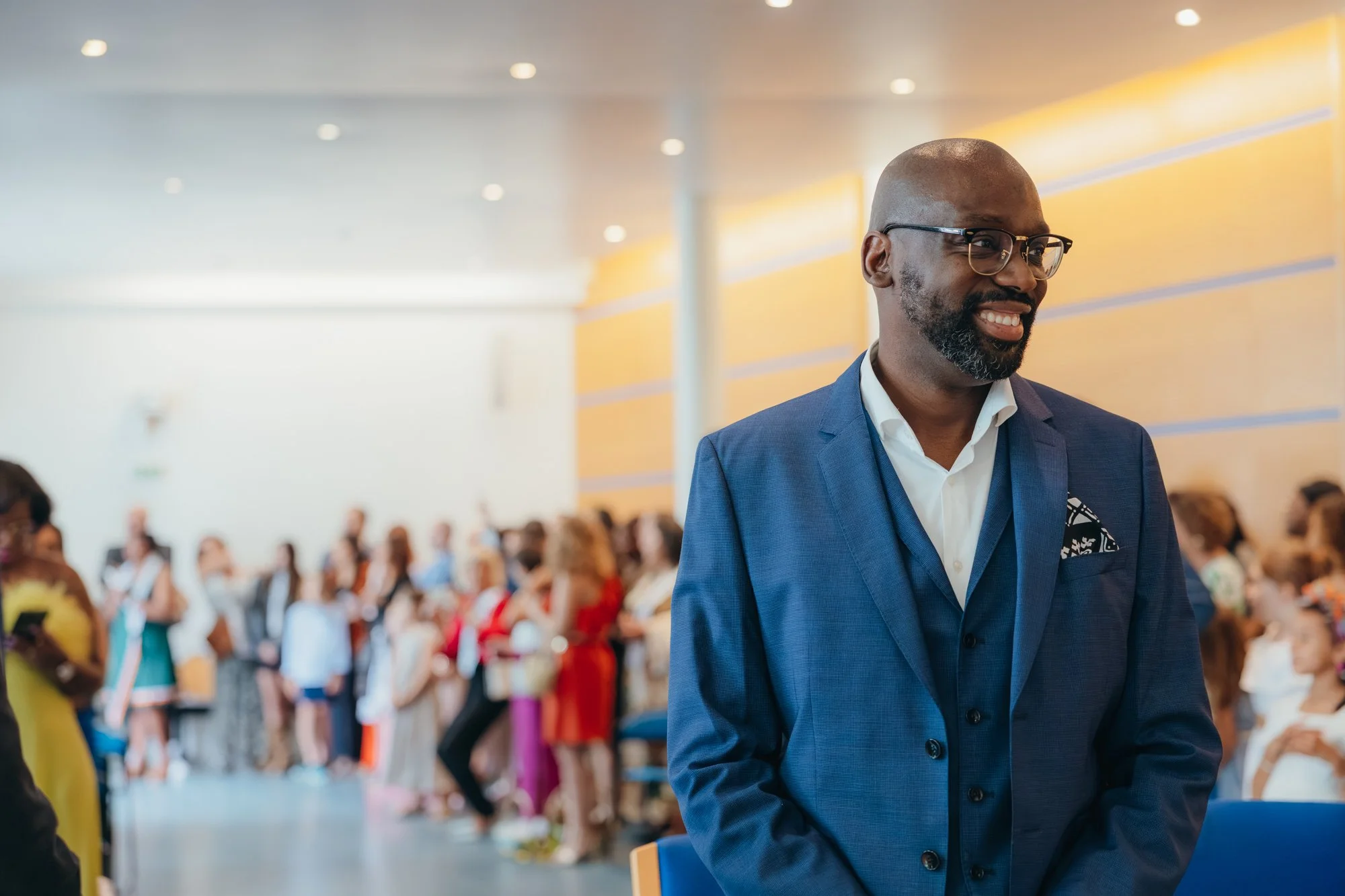 Un homme souriant en costume bleu, portant des lunettes, lors d'un événement professionnel avec une foule en arrière-plan dans une salle bien éclairée.