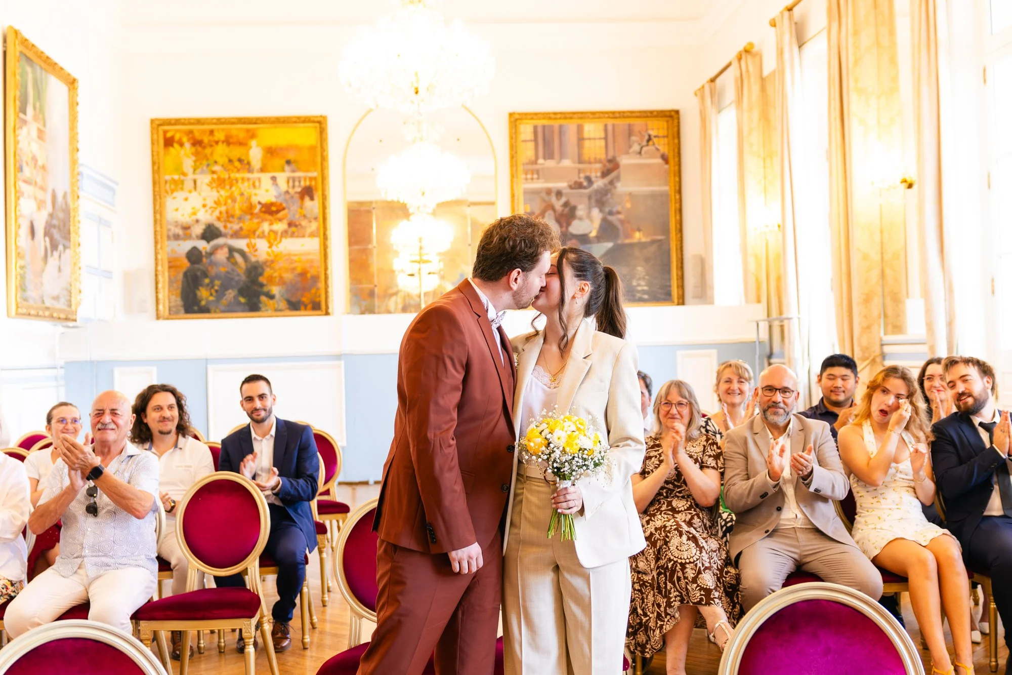Un couple de mariés s'embrasse lors de leur mariage en présence d'une assemblée applaudit, dans une salle ornée de tableaux et luminaires chandeliers, avec des invités souriants et habillés élégamment.