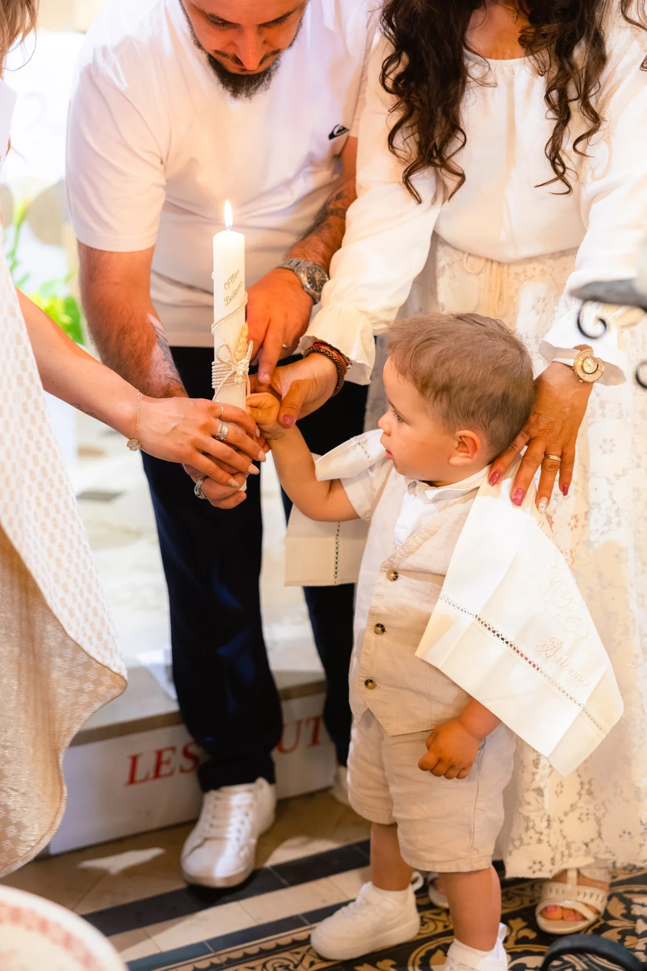Un jeune garçon participe à une cérémonie religieuse, aidé par des adultes pour allumer une bougie lors d'un rituel, vêtus de vêtements légers et blancs dans un lieu intérieur.»