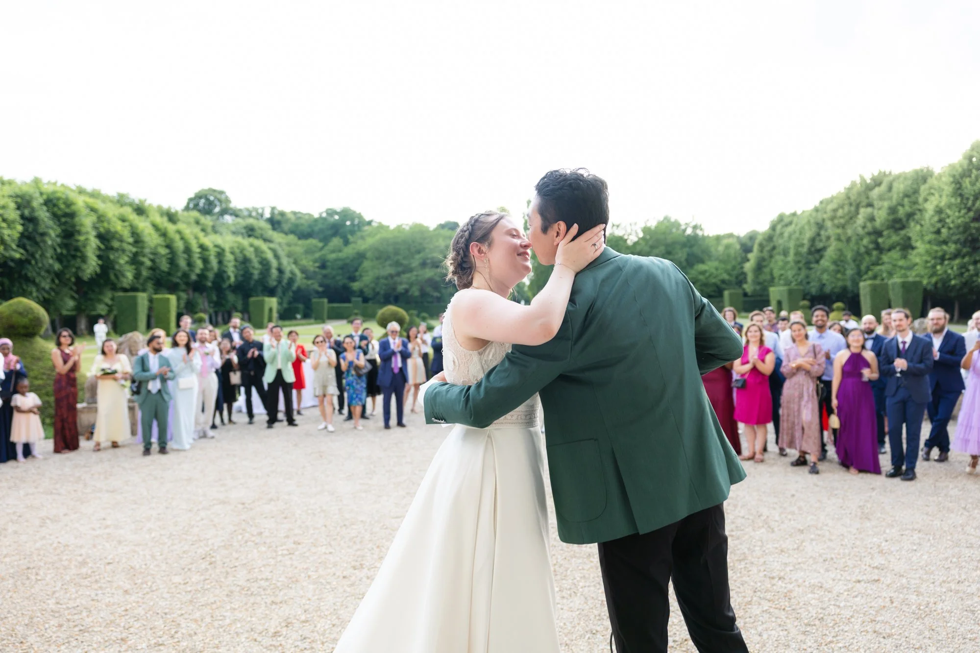 Un couple en robe de mariée et costume, s'embrassant lors d'une cérémonie en plein air, avec une foule d'invités debout derrière eux, dans un jardin élégant.