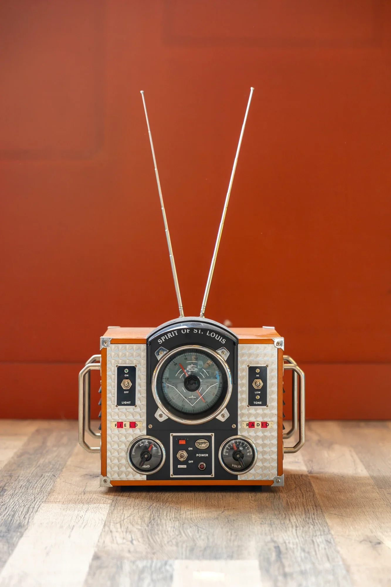 Une vieille radio avec des antennes en V, placé sur un sol en bois, contre un mur rouge-orange.