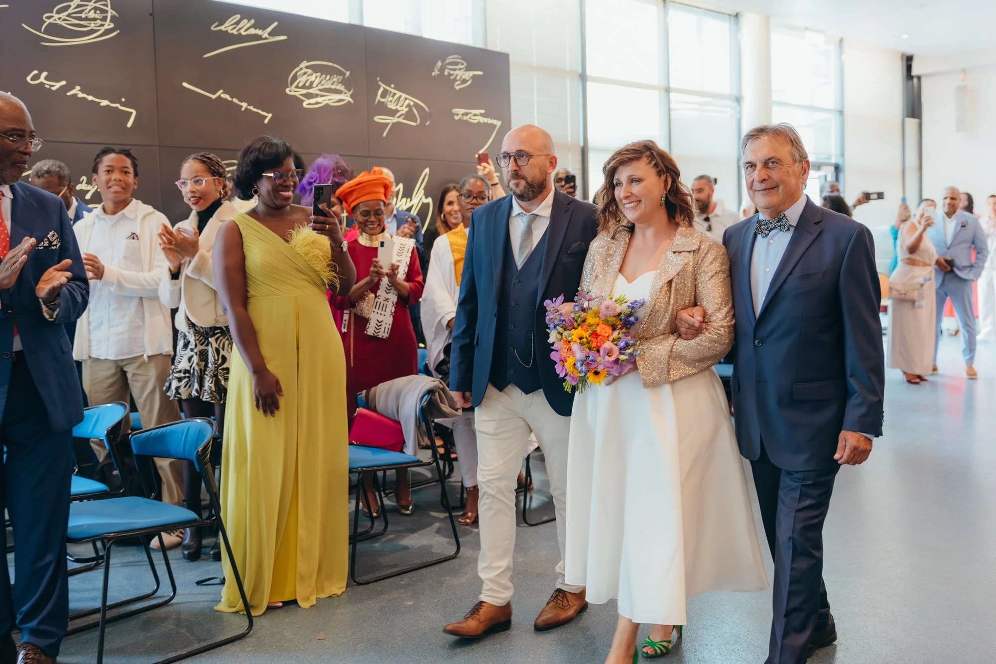 Groupe de personnes lors d'une cérémonie ou d'une célébration dans un lieu moderne avec des grandes fenêtres, concentrées autour d'une femme tenant un bouquet de fleurs et accompagnée d'un homme. L'ambiance est joyeuse et festive.