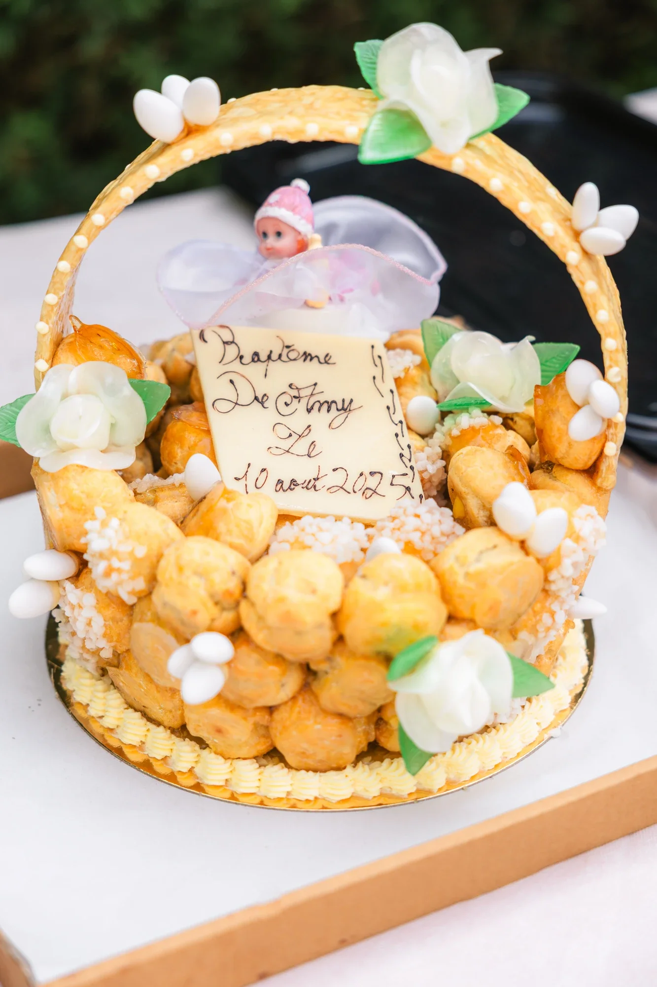 Gâteau de baptême décoré avec des fleurs en pâte d'amande, une figurine de bébé, et une plaque d'inscription en français.