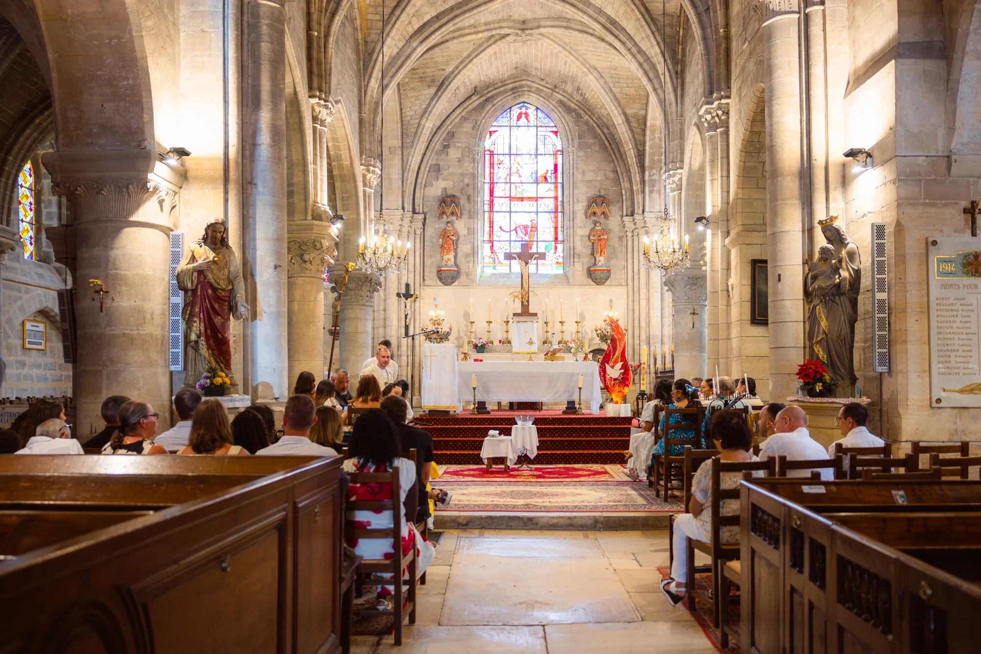 Intérieur d'une église gothique avec des vitraux colorés, des statues religieuses et un autel au centre. La salle est remplie de personnes assises, probablement pour une cérémonie religieuse.