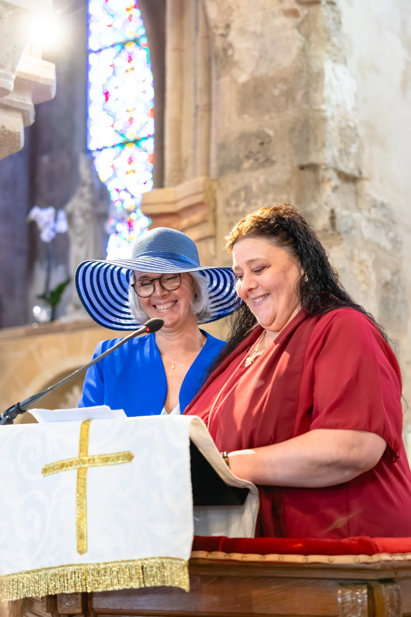 Deux femmes souriantes, l'une portant un chapeau à rayures bleues et blanches et des lunettes, l'autre avec des cheveux noirs, semblent être lors d'une cérémonie ou d'un événement religieux dans un lieu en pierre avec des vitraux colorés en arrière-p