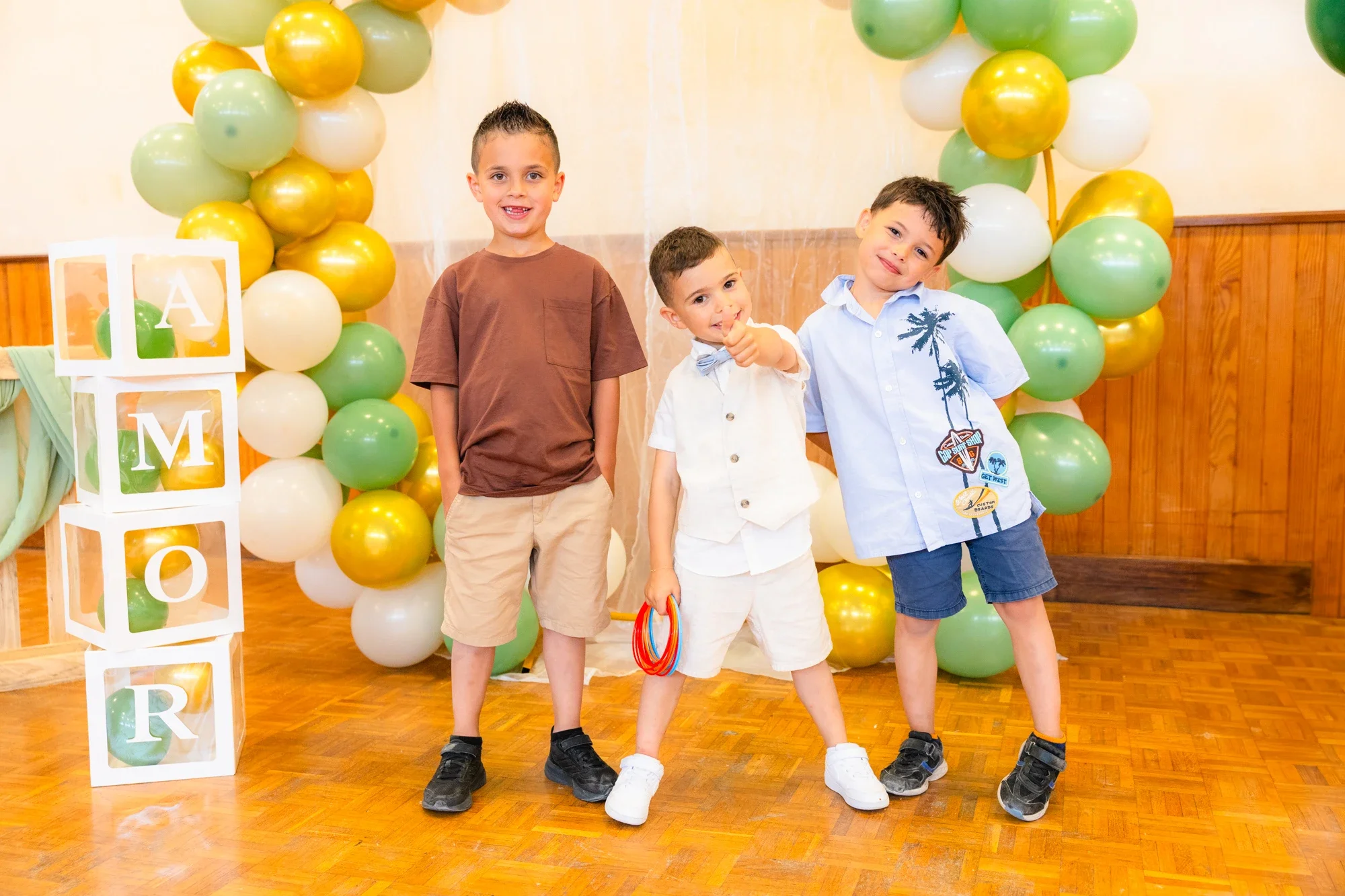 Trois garçons posant devant une décoration de ballons verts, jaunes, blancs et dorés, avec des blocs en carton blanc portant des lettres et un ballon en forme de cercle multicolore.