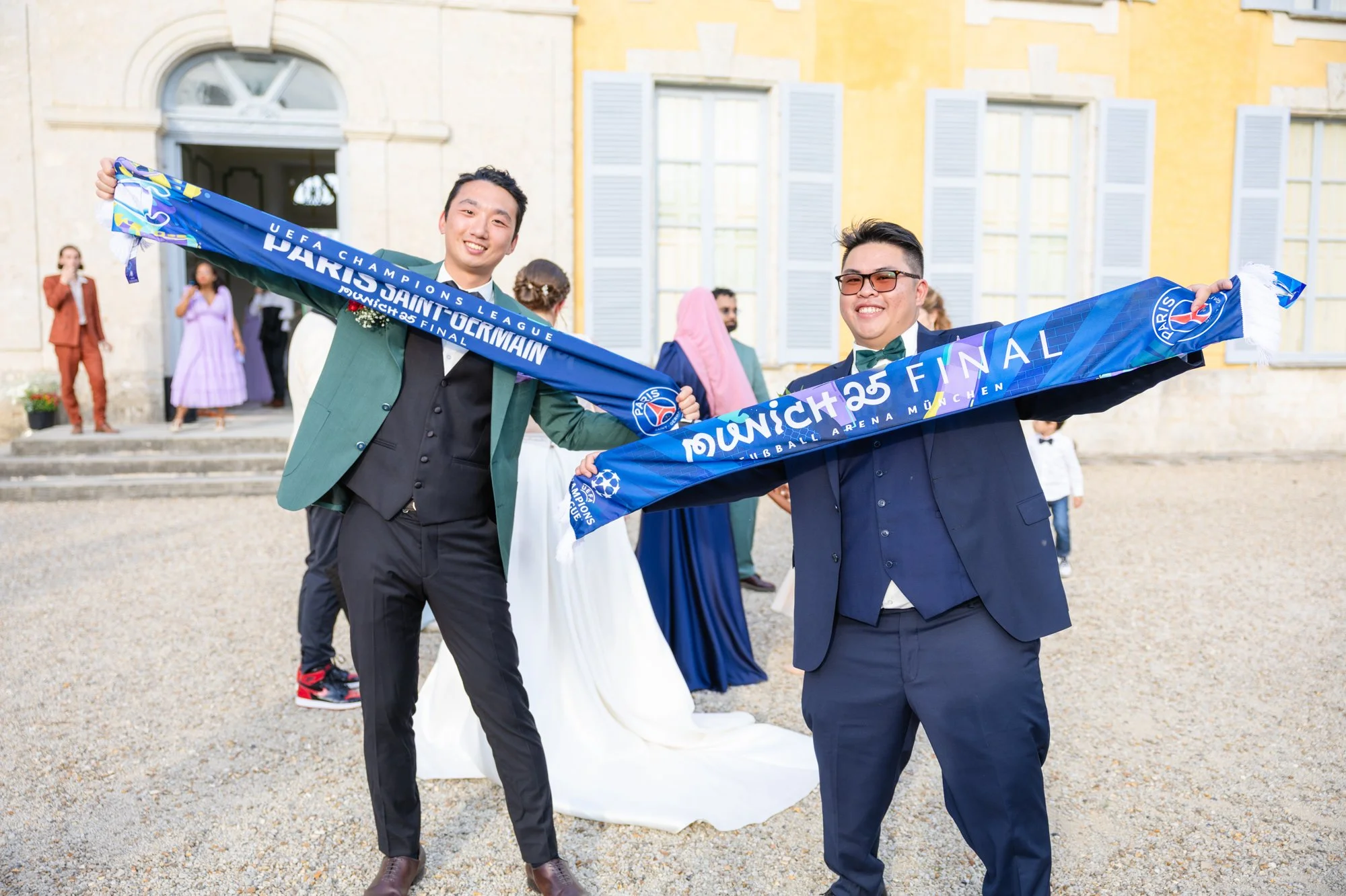 Deux jeunes hommes habillés en costume sont souriants et tiennent une écharpe en satin bleu avec le texte 'UEFA Champions League Paris Saint-Germain Match 25 Final.' Ils se tiennent devant une maison de style ancien avec des volets bleus, lors d'une 