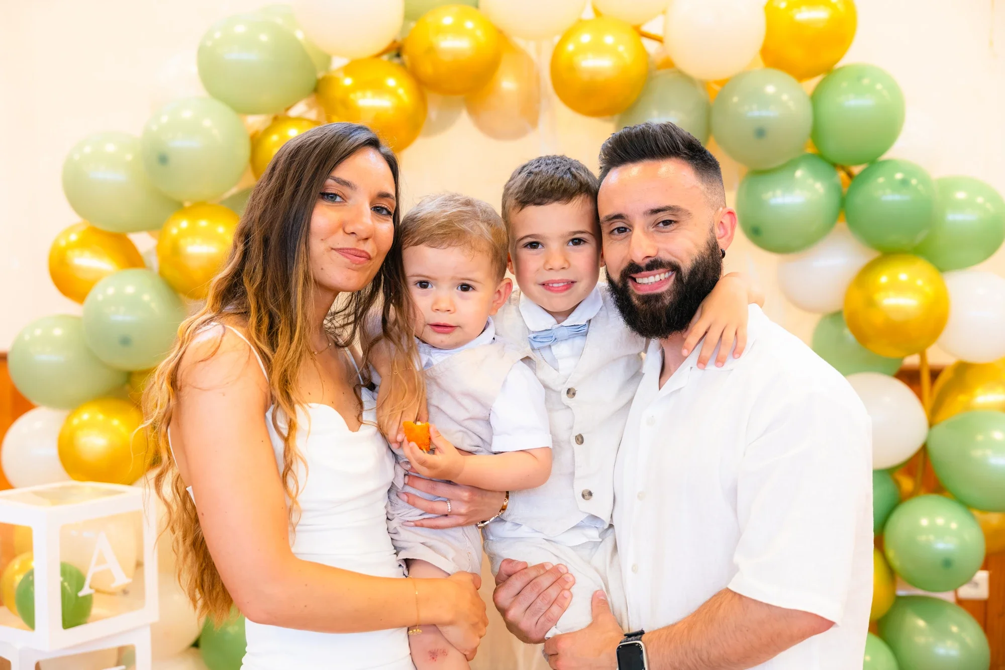 Une famille de quatre personnes posant devant un décor de ballons verts, jaunes et blancs lors d'une fête d'anniversaire, avec un gâteau en arrière-plan.