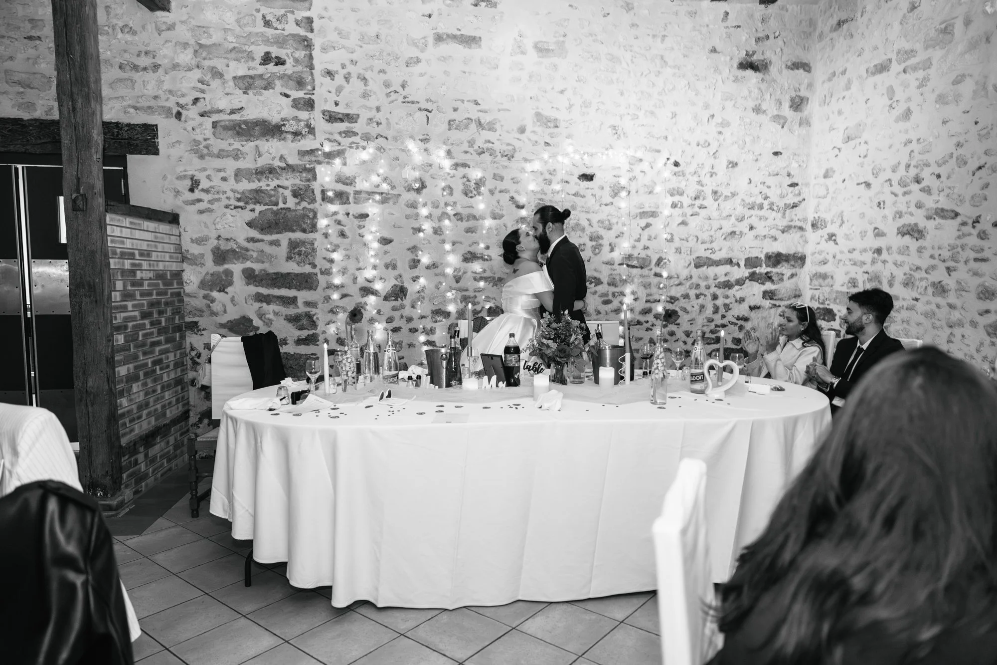 Un couple de mariés s'embrasse au centre d'une table décorée lors d'une réception de mariage, avec des invités assis autour.
