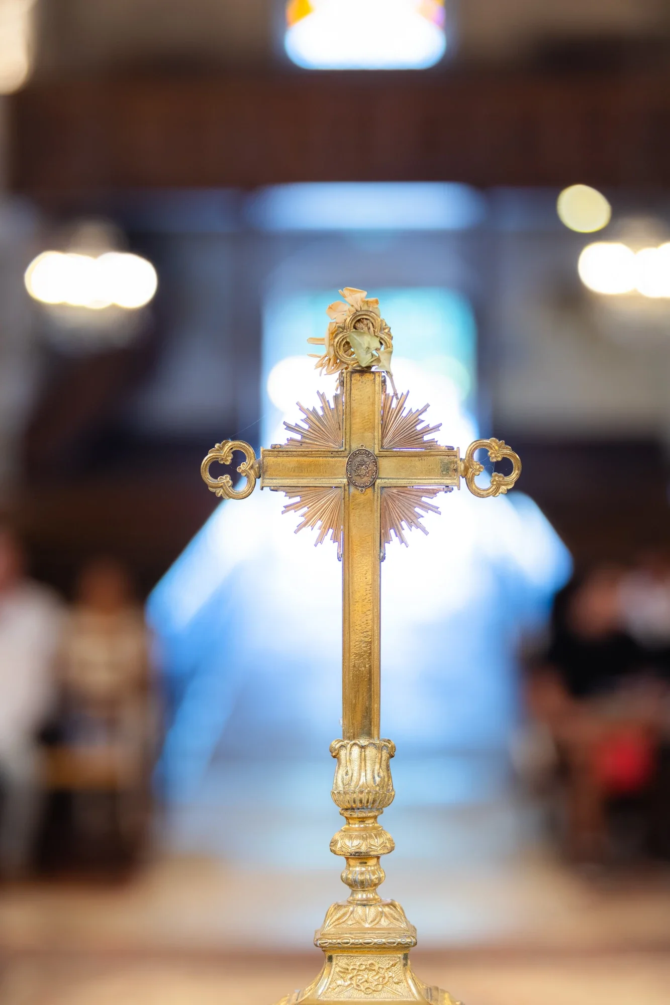 Une croix en or ornée d'une couronne et d'une figurine, placée sur un socle décoratif, dans un espace intérieur avec un fond flou