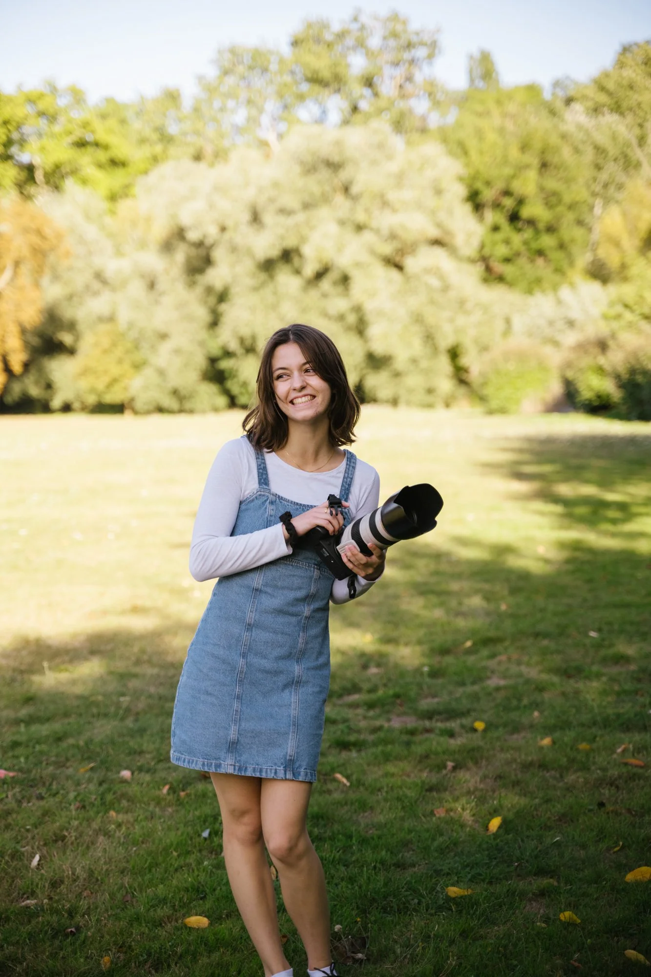 Une jeune femme avec des cheveux bruns portant une robe en jean et une chemise blanche, tenant un grand téléobjectif, se tient dans un parc ensoleillé avec des arbres verts en arrière-plan.