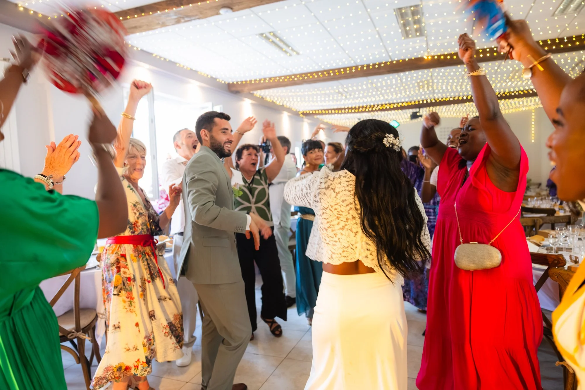 Des personnes dansent et s'amusent lors d'une fête dans une salle décorée avec des lumières suspendues. La scène est joyeuse avec des gens de divers horizons qui sourient et lèvent les bras.
