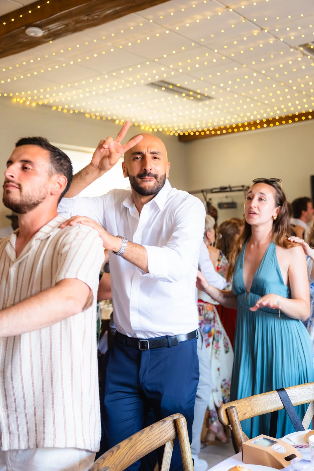 Groupe de personnes lors d'une fête, avec un homme au centre faisant un geste de paix derrière une autre personne. Décor avec des lumières suspendues au plafond.