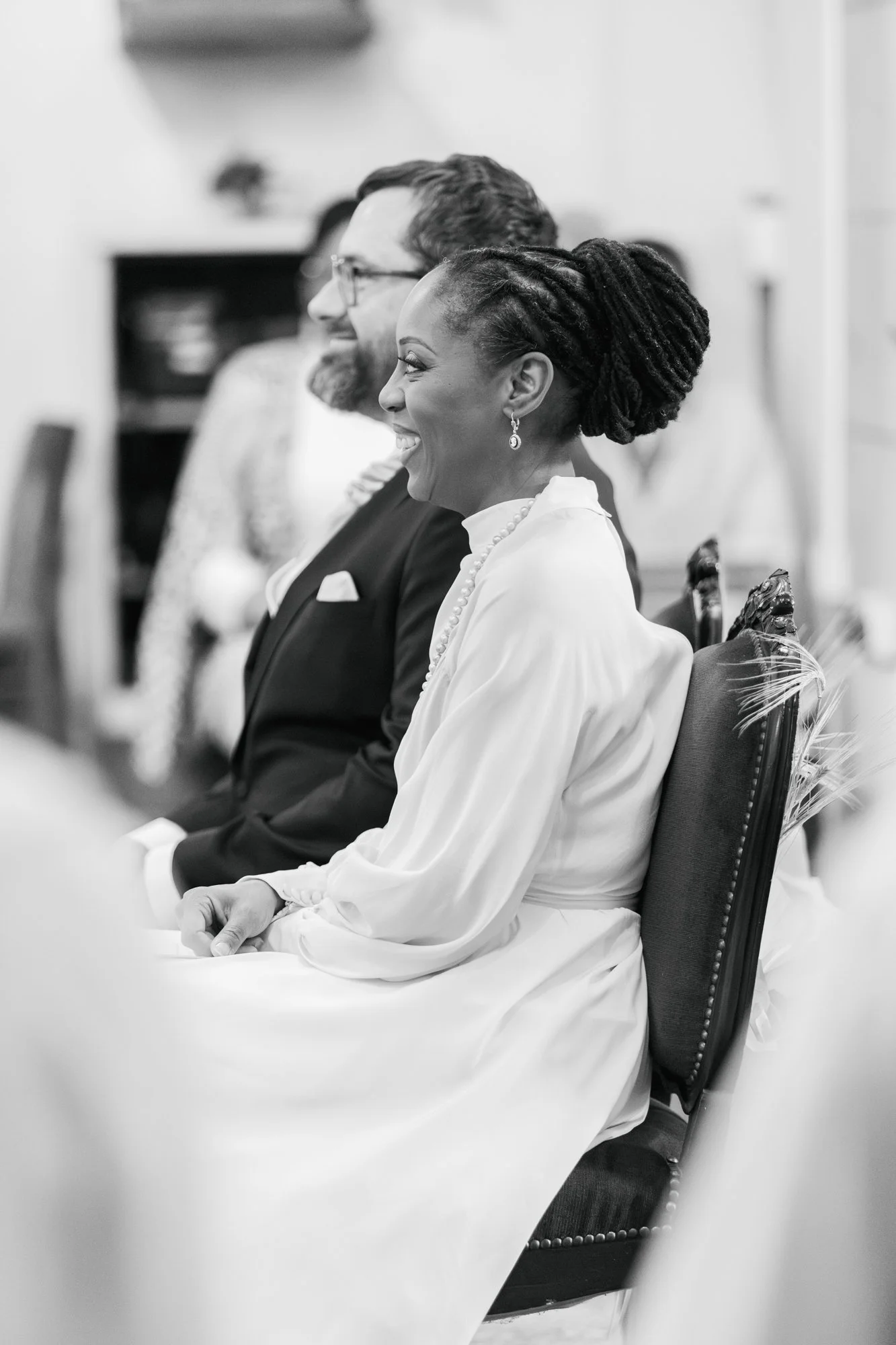 Deux personnes assises côte à côte lors d'une réunion ou d'un événement. La femme porte une robe blanche, des boucles d'oreilles pendantes et un collier, et a ses cheveux en dreadlocks. L'homme porte un costume sombre avec une chemise blanche. Ils so