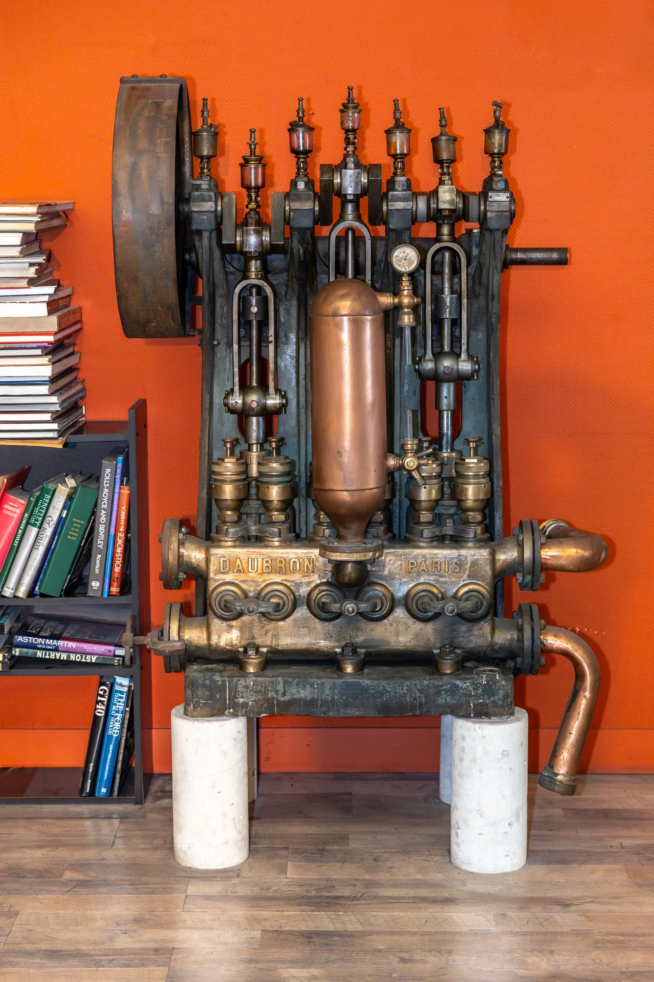 Machine ancienne en métal avec des tubes, des jauges et un grand volant, posée sur deux colonnes en pierre, avec une étagère de livres à gauche et un mur orange en arrière-plan.