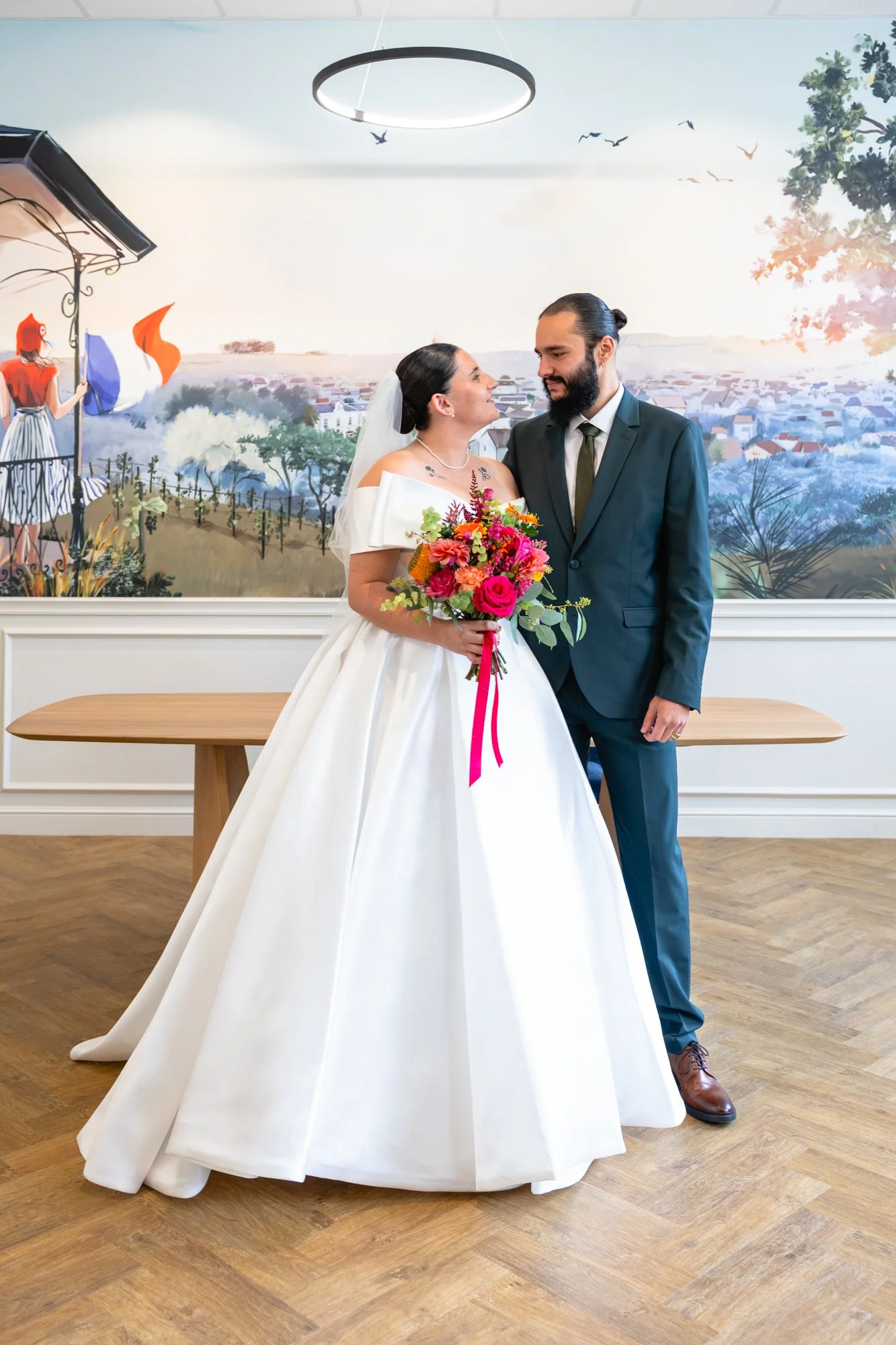 Un couple en costume de mariage, la femme tenant un bouquet de fleurs, se tenant face à face devant une peinture murale représentant un coucher de soleil sur une ville.