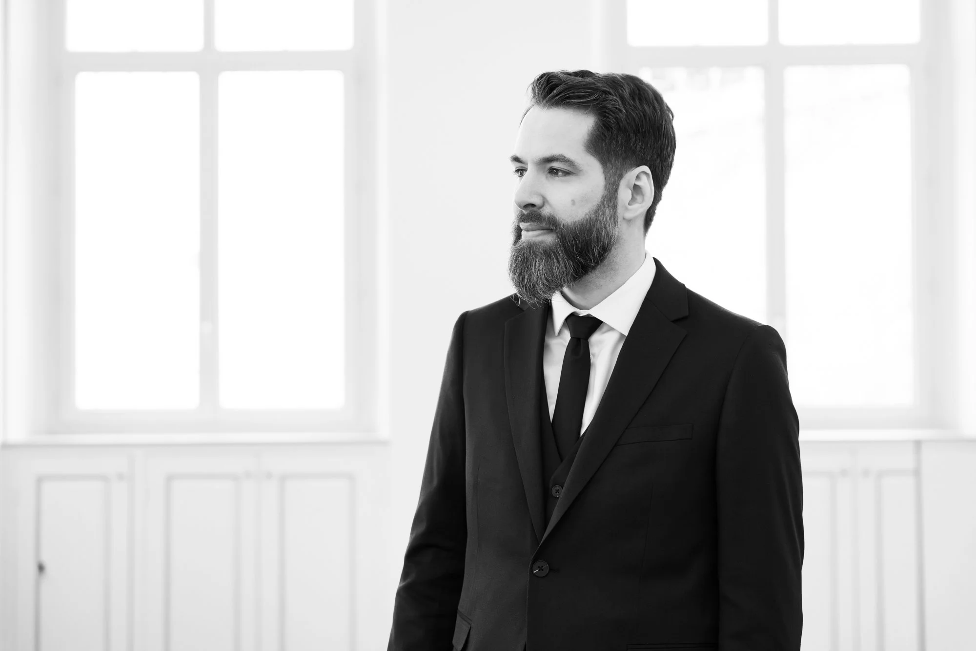 Un homme en costume noir avec une barbe regarde vers la droite dans une pièce lumineuse avec de grandes fenêtres.