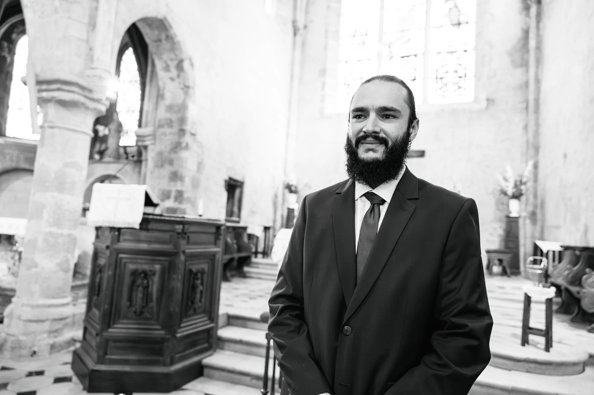 Homme en costume dans une église avec des piliers en pierre et des vitraux.