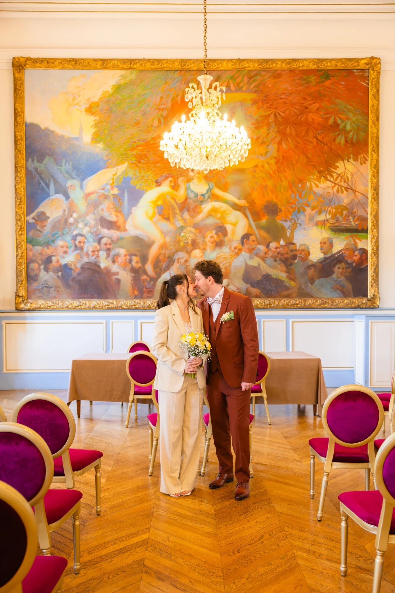 Un couple habillé élégamment, probablement lors d'un mariage, se tient dans une salle ornée d'un grand tableau coloré représentant une scène artistique et d'un lustre chandelier.'