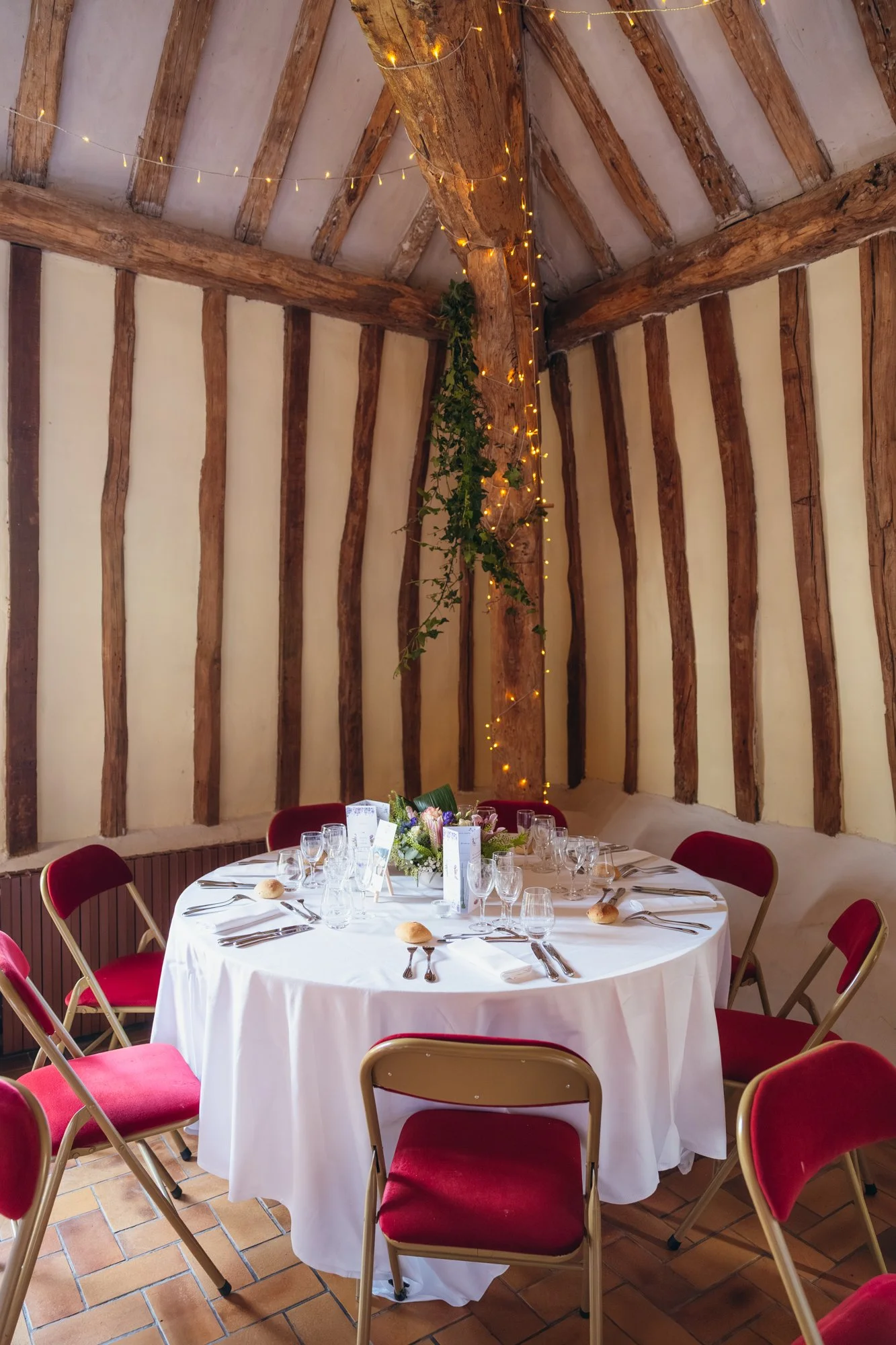 Une table dressée pour un repas avec des bougies, des verres, des assiettes et un bouquet de fleurs, dans une pièce avec des murs en bois et une décoration de lumières