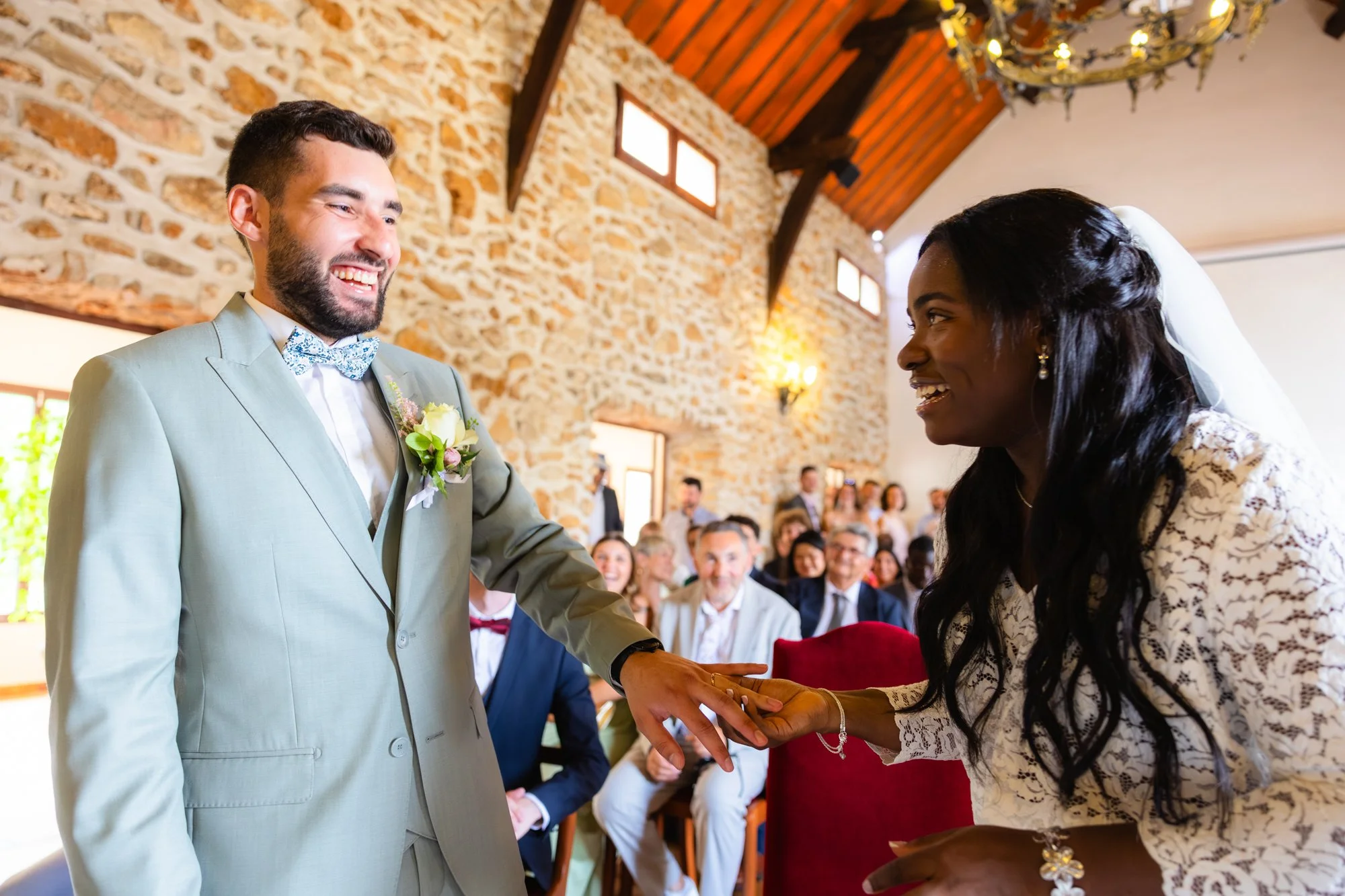 Un couple lors d'une cérémonie de mariage, l'homme en costume gris et la femme en robe de mariage, échangeant un anneau dans une salle avec murs en pierre et plafond en bois, entourés de nombreux invités.