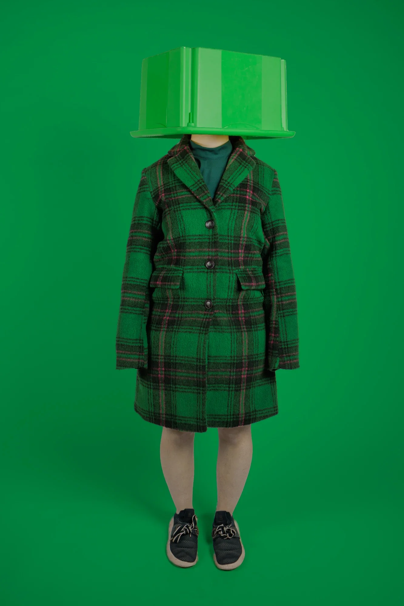 Une personne habillée d'un manteau à carreaux vert et rouge, avec un sac en plastique vert sur la tête, posant devant un fond vert.