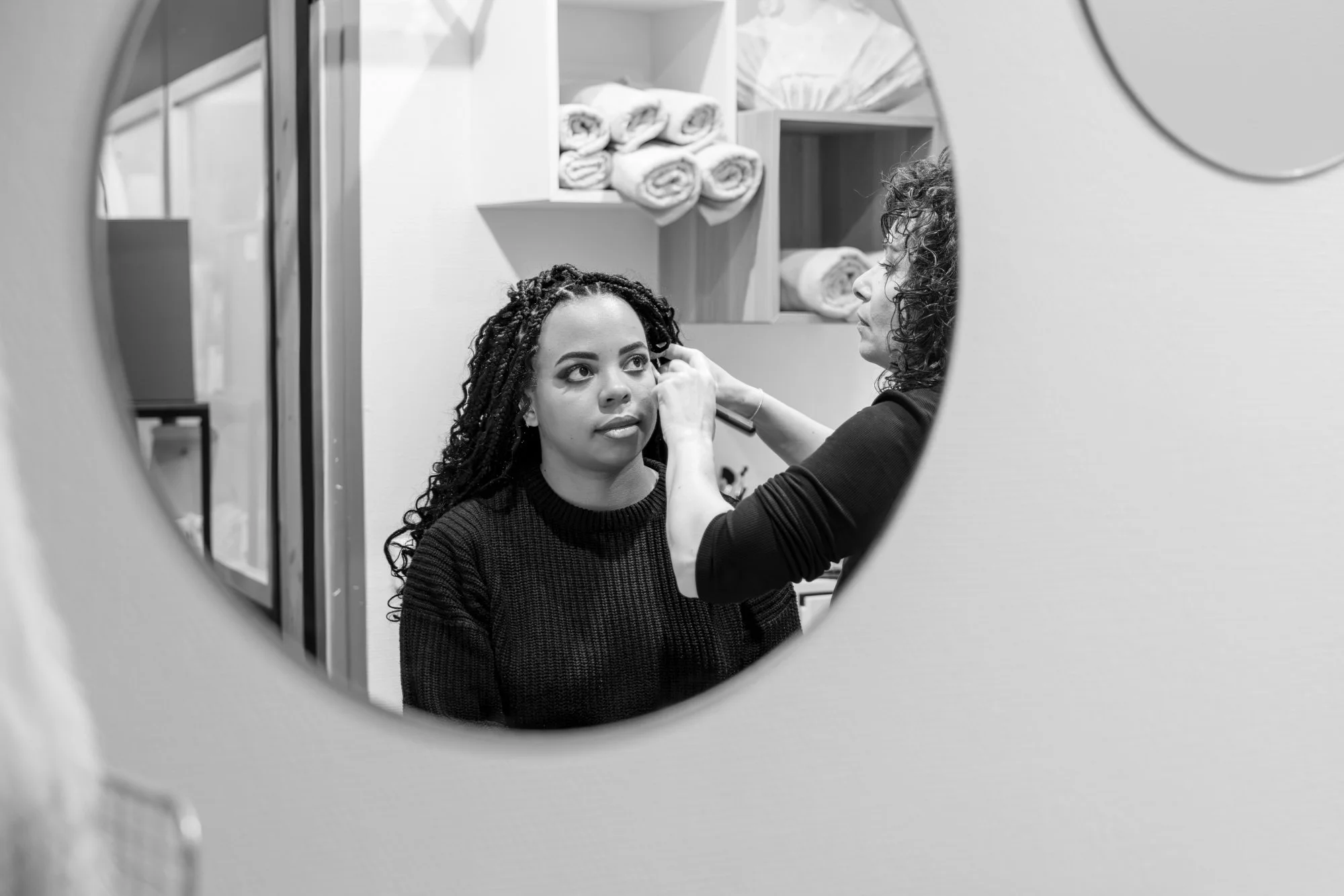 Une femme regarde dans un miroir rond, une autre femme ajuste ses boucles d'oreilles, derrière un mur avec des étagères contenant des serviettes roulées.