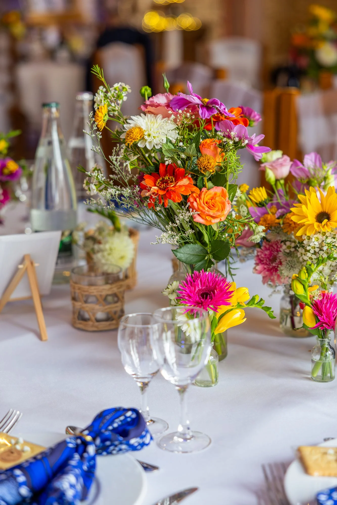 Un centre de table avec un arrangement floral coloré sur une table recouverte d'une nappe blanche lors d'une réception ou d'un repas. Parmi les fleurs, on distingue des roses, des tournesols et d'autres fleurs multicolores.