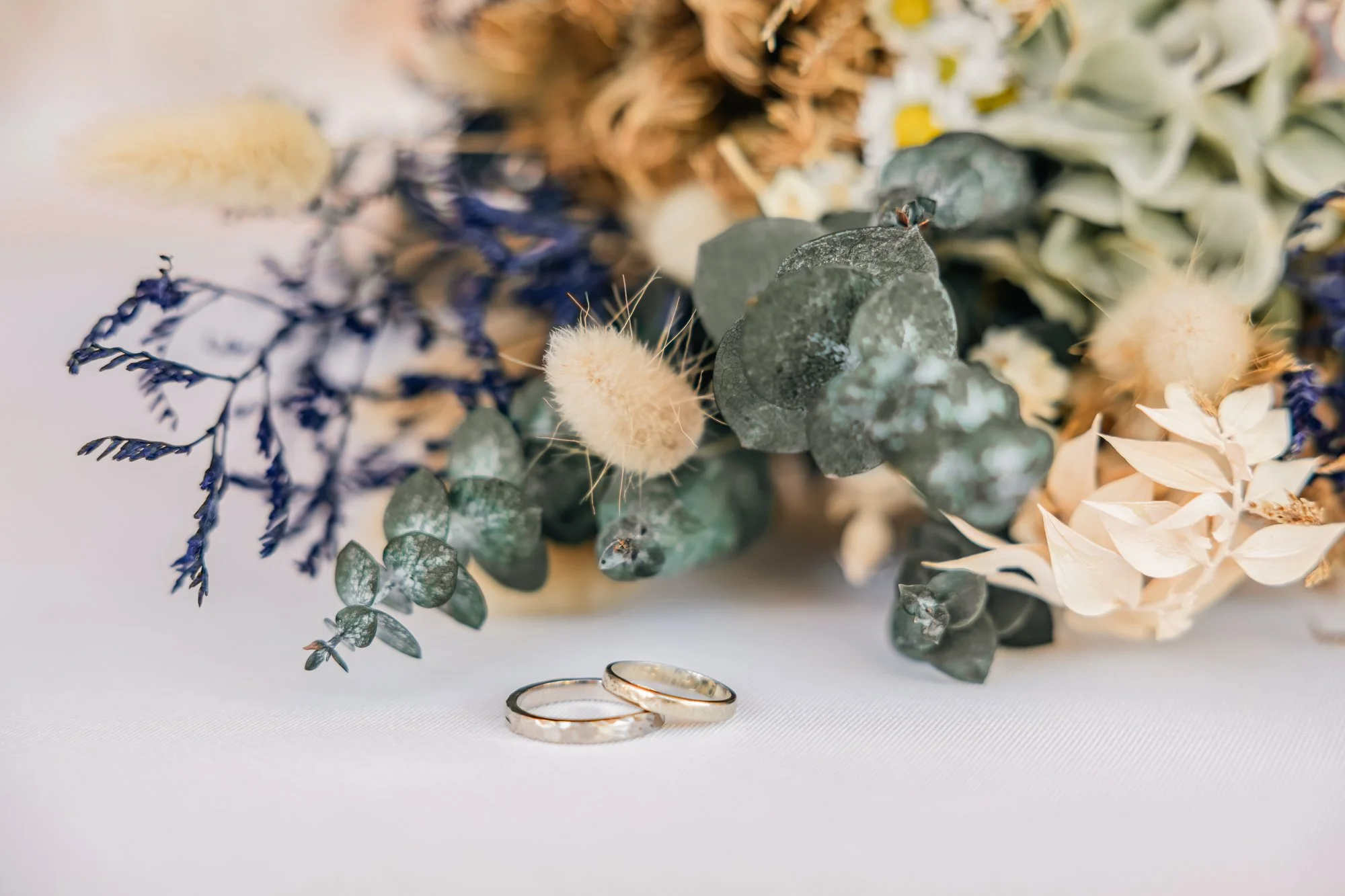 Un bouquet de fleurs séchées avec deux alliances en or posées devant. La composition comprend des eucalyptus, des fleurs violettes, et des éléments doux comme des fleurs blanches et des peluches écuyères.