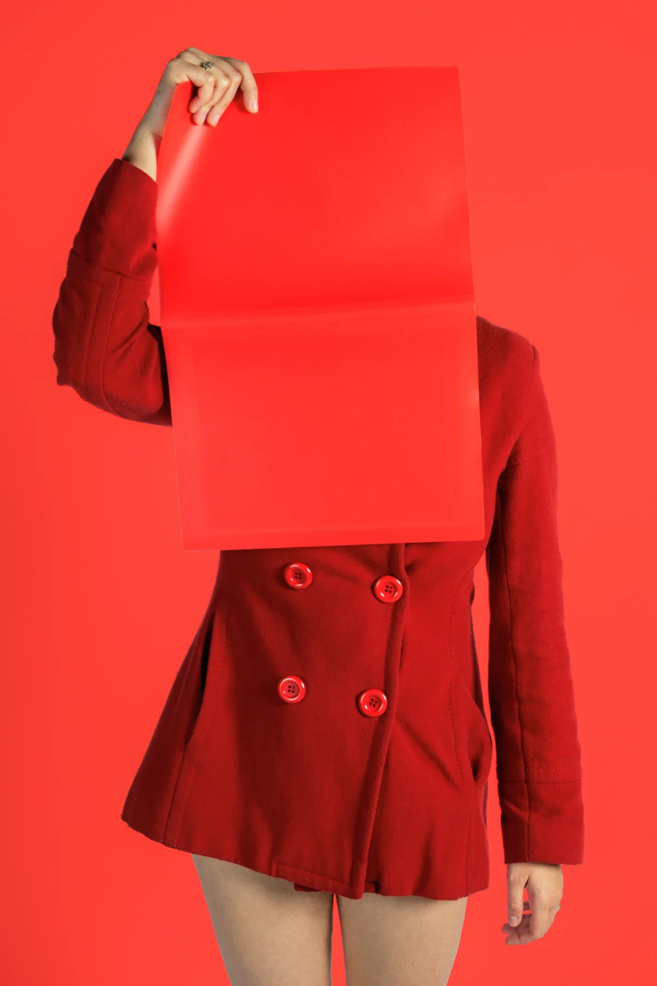 Une personne porte un manteau rouge avec des boutons, tenant une plaque rouge devant leur visage, avec un fond rouge uniforme.