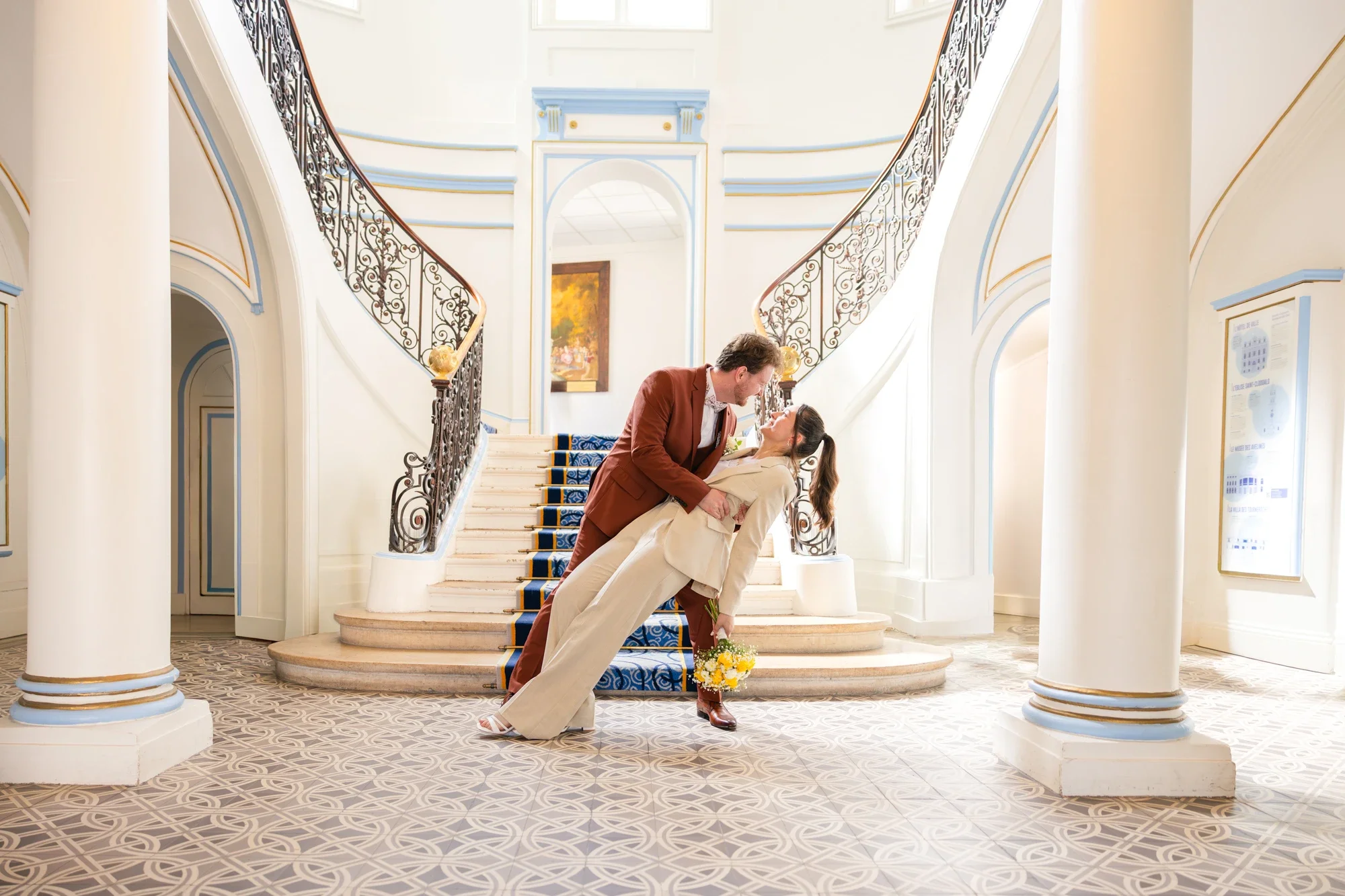 Un couple dans une scène romantique sur un escalier en marbre blanc avec une rampe en fer forgé. La femme est penchée en arrière, tenant un bouquet de fleurs jaunes, tandis que l'homme se penche vers elle, souriant. La scène se déroule dans un intérieur lumineux avec des colonnes blanches et des murs ornés de moulures bleues et dorées, et une peinture derrière eux.