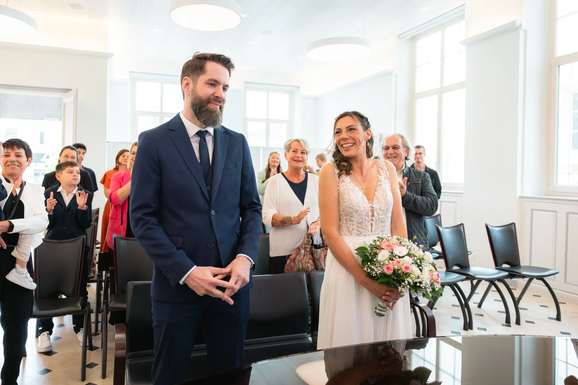 Un couple de mariés lors d'une cérémonie de mariage, entouré de proches applaudissant et souriant dans une salle lumineuse.