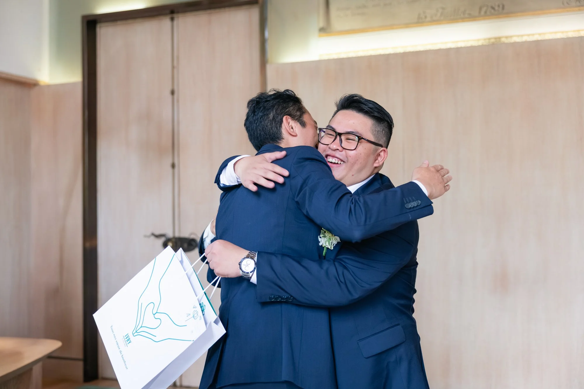 Deux hommes habillés en costume, l'un avec des lunettes, s'embrassent dans une accolade lors d'un événement en intérieur.