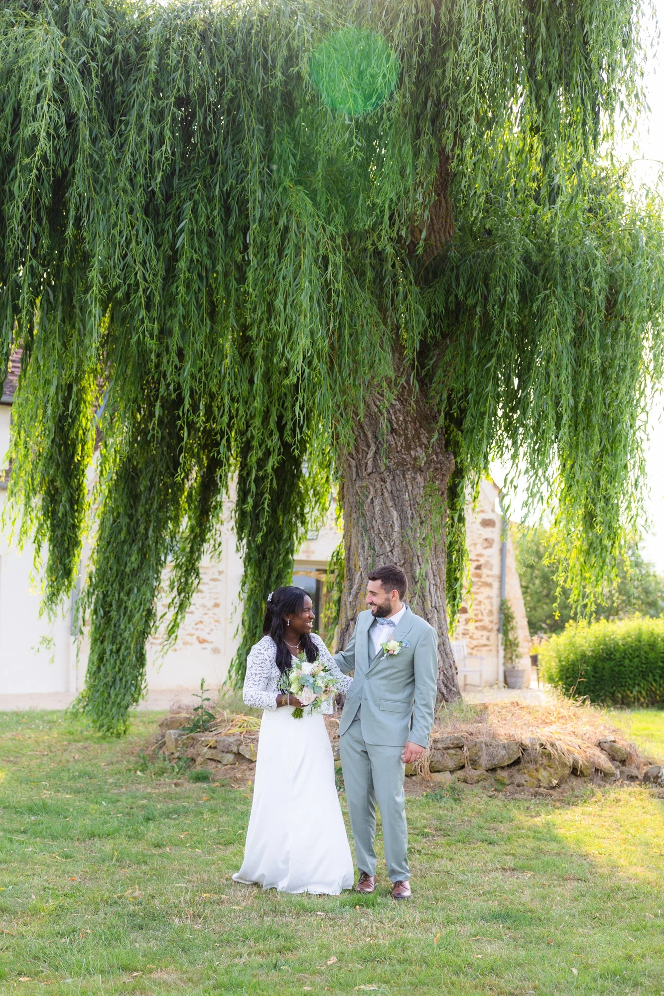 Un couple lors d'un mariage en plein air sous un arbre à feuilles longues et suspendues, se tenant par la main et échangeant un regard, avec une maison en pierre en arrière-plan.