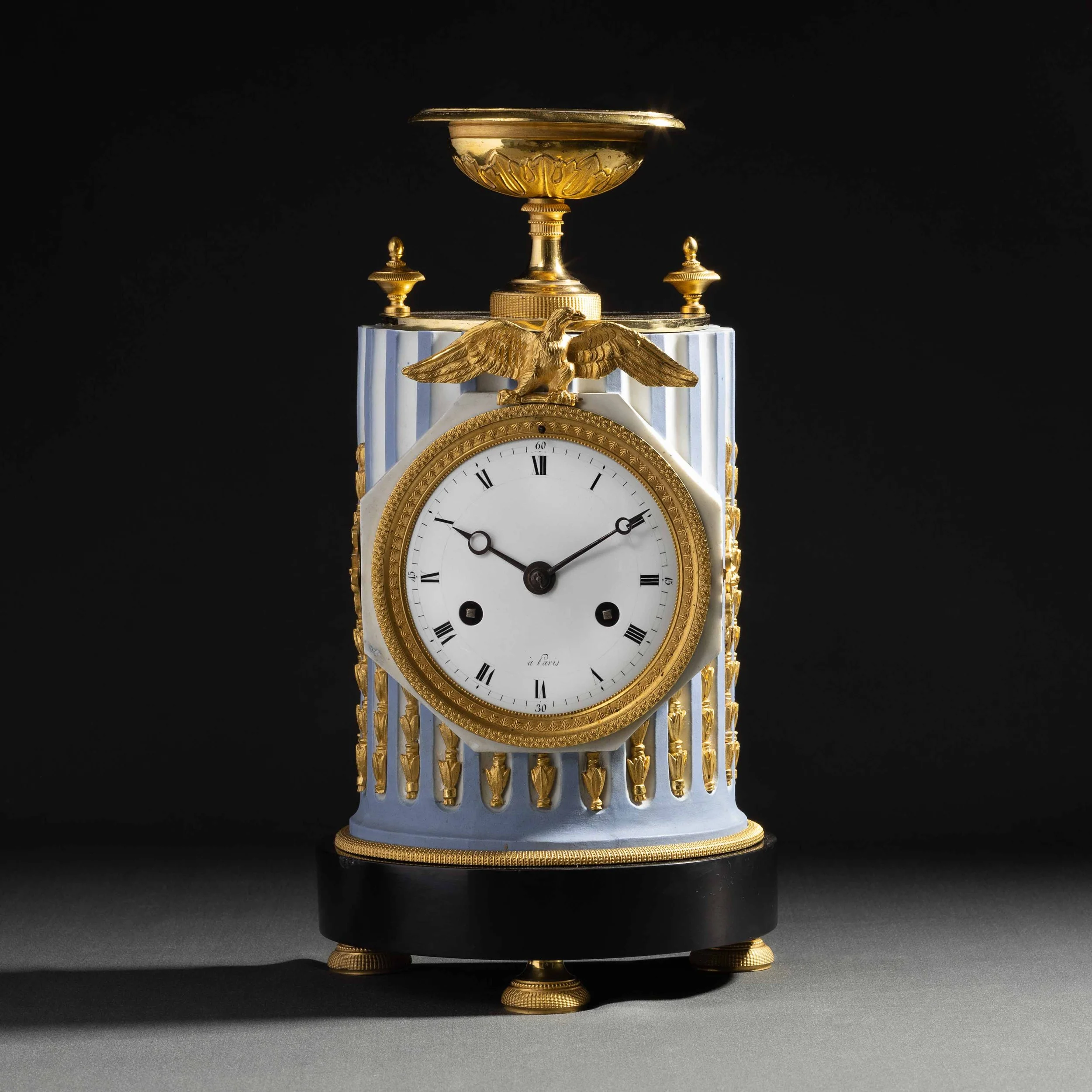Une horloge décorative avec une structure en or blanc et or jaune, surmontée de deux petites tours et d’un aigle en or en relief. La partie visible de la face de l’horloge affiche des chiffres romains et indique 2h10.