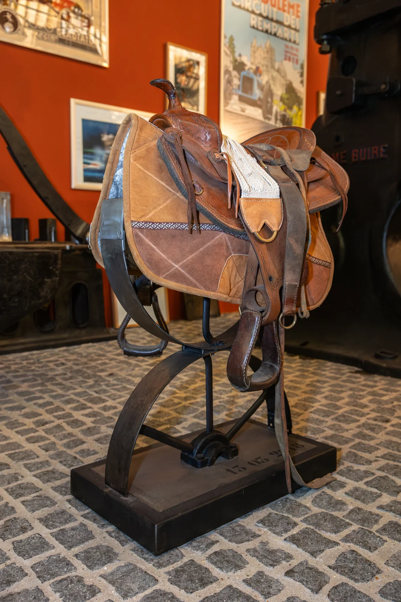 Une selle de cheval ancienne exposée sur un support en musée, avec des affiches et des œuvres d'art en arrière-plan.