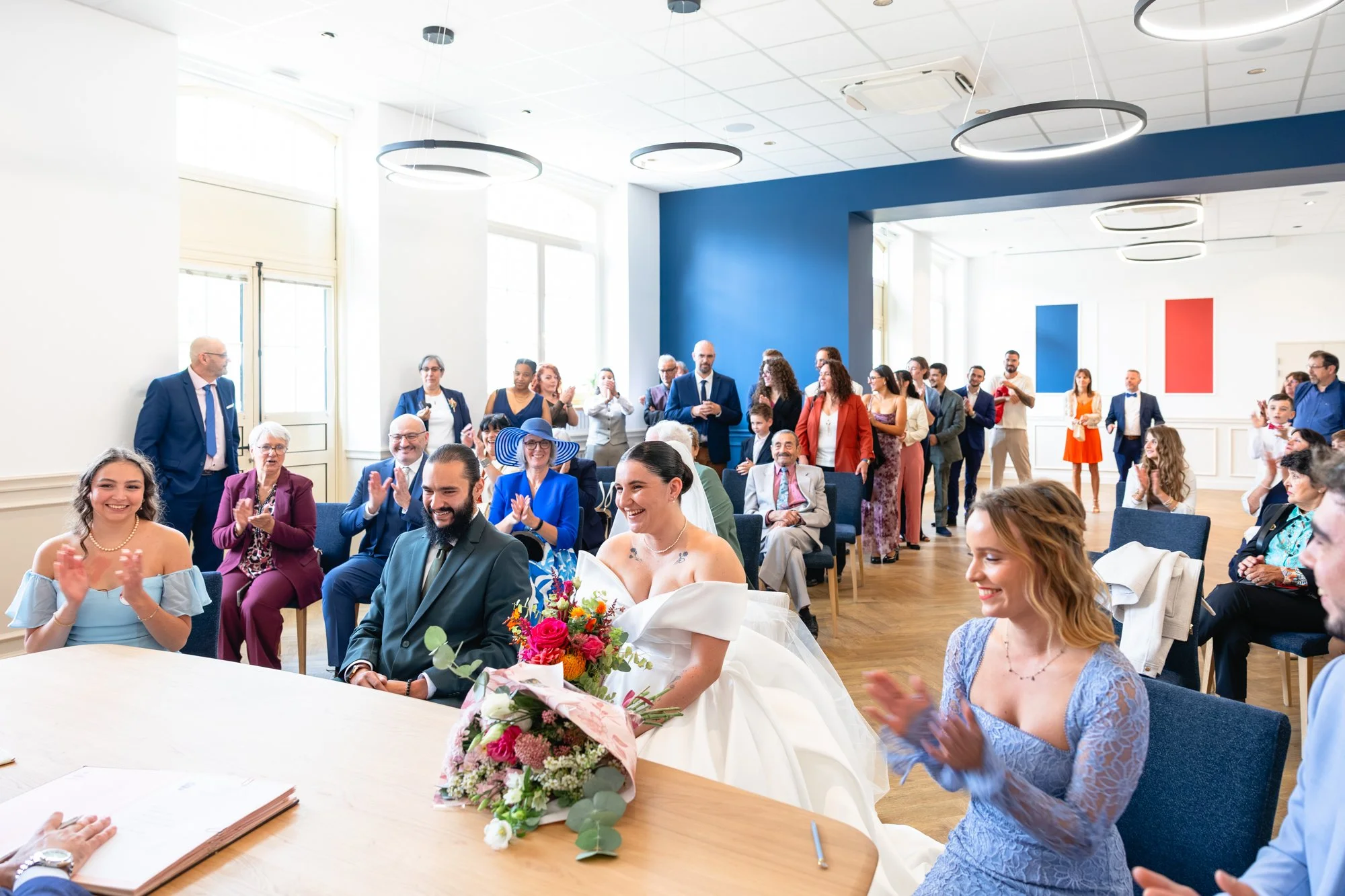 Un groupe de personnes lors d'une cérémonie de mariage, avec la mariée devant une bouquet de fleurs, des invités qui applaudissent et un espace intérieur décoré en blanc et bleu.