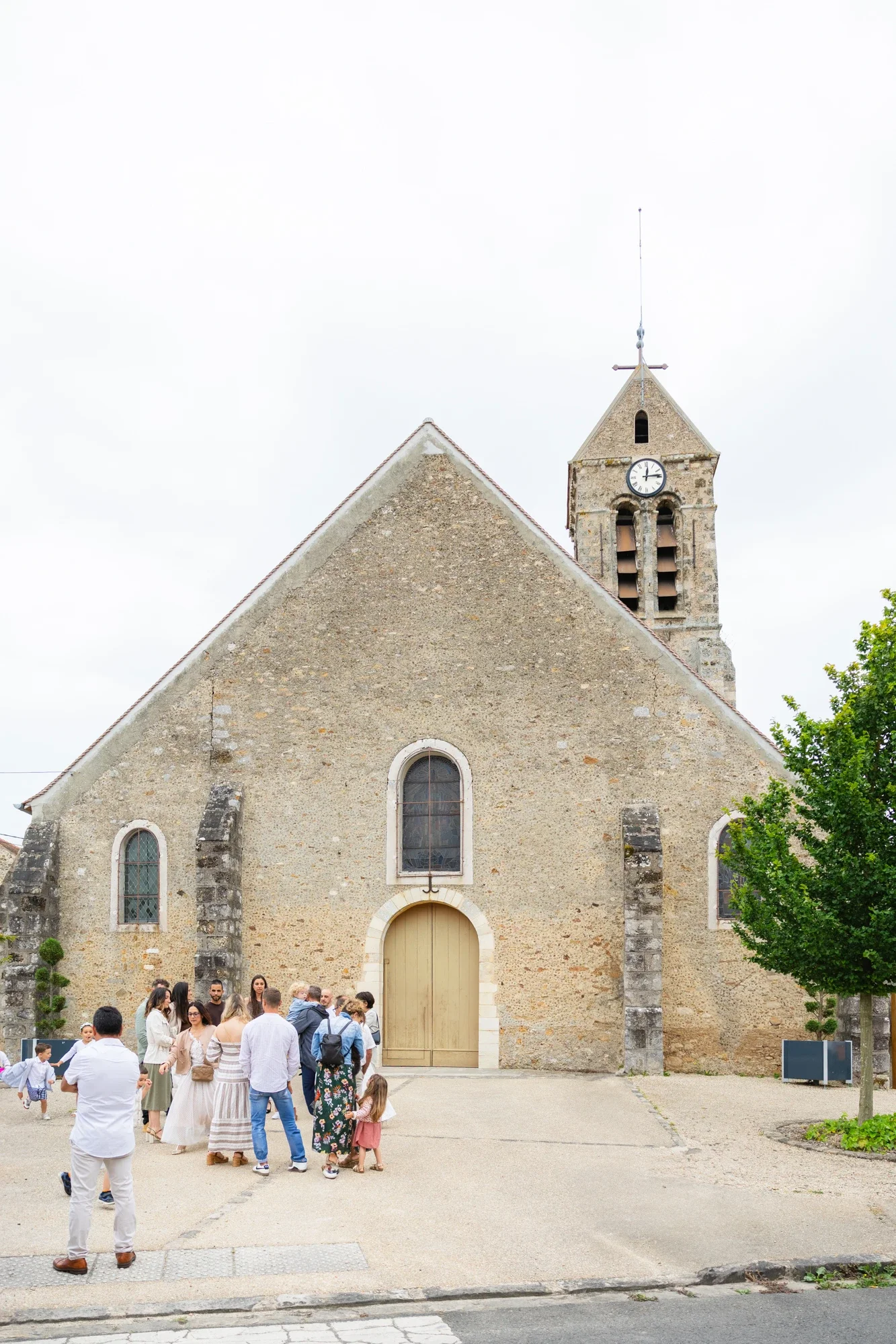 Une église ancienne en pierre avec une horloge sur la tour, entourée de plusieurs personnes, certaines attendant devant l'entrée, dans un endroit public. Il y a aussi un arbre vert à droite.